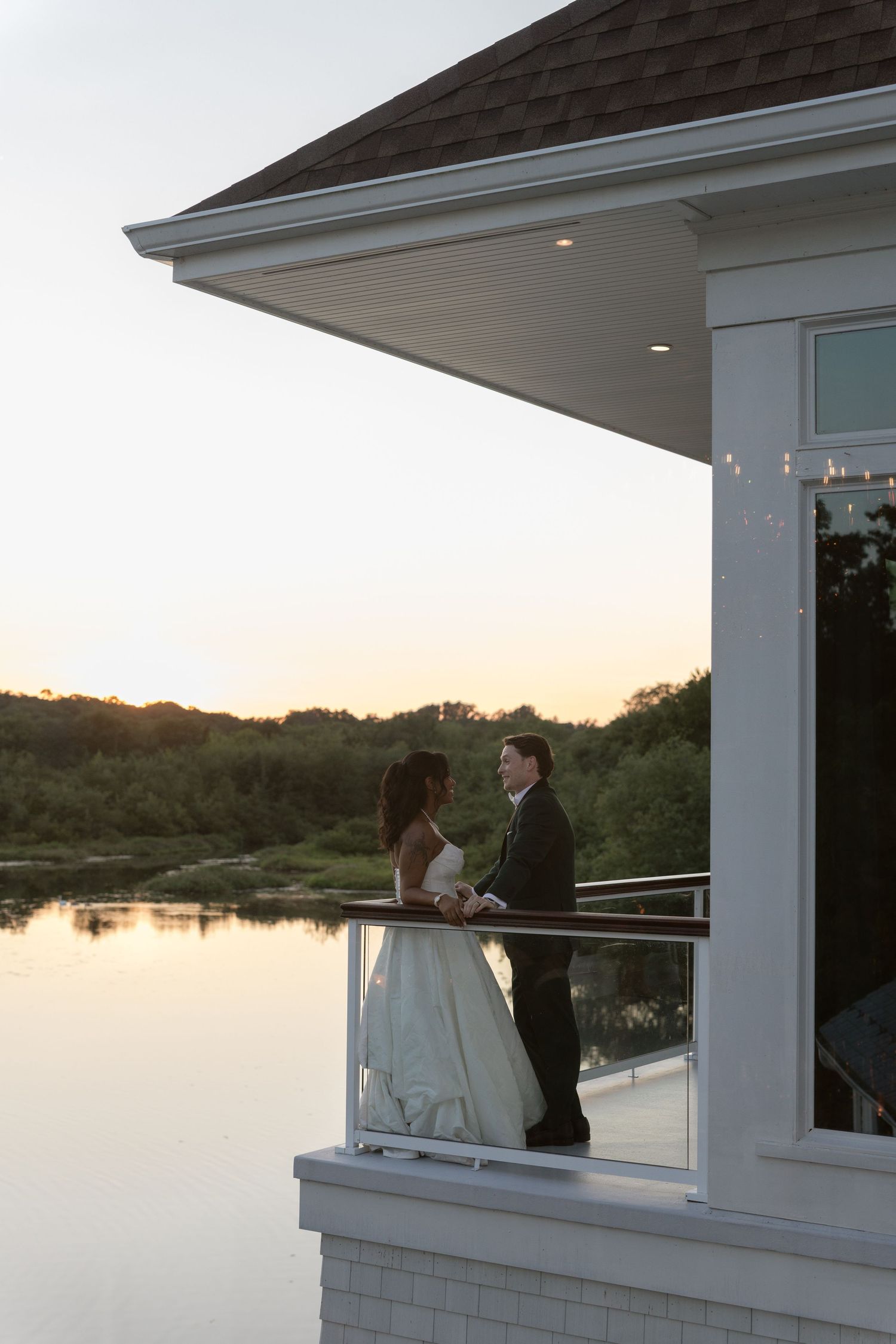 Bride and groom share a romantic moment on a balcony overlooking a serene lake at sunset.