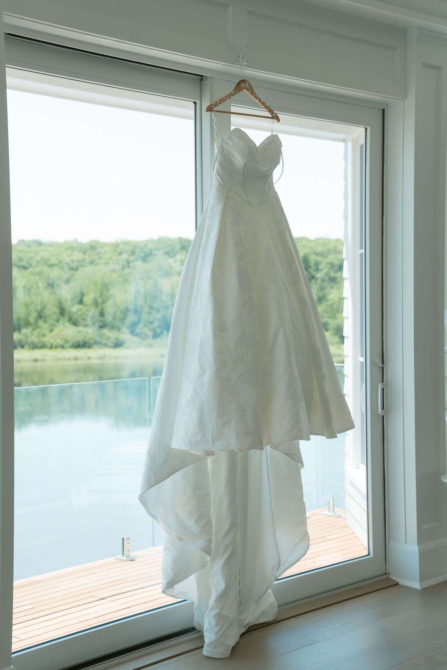Elegant strapless white ball gown wedding dress hanging on a hanger in front of a large window overlooking a serene lake.