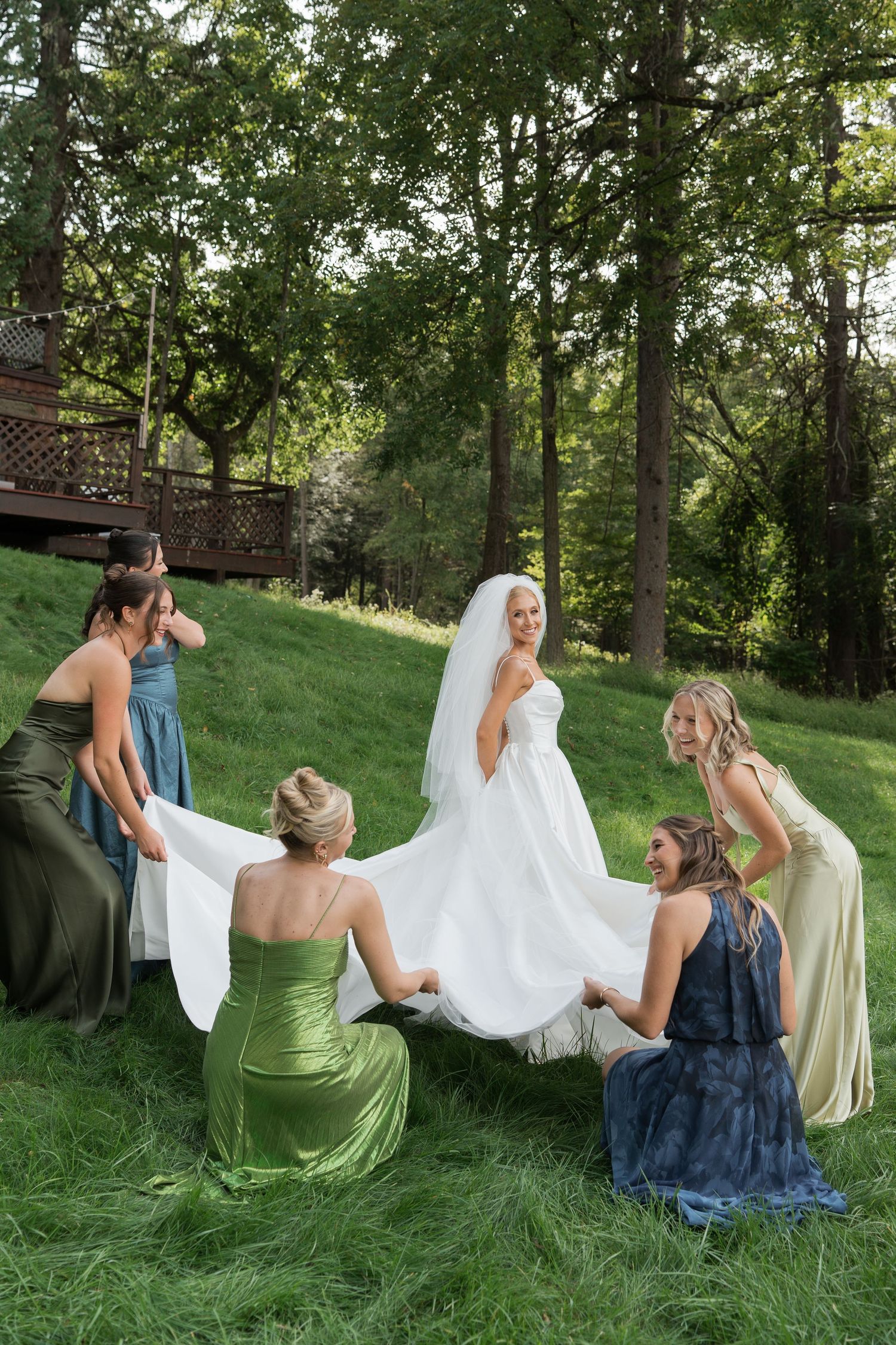 Bride in white gown and veil surrounded by bridesmaids in colorful dresses arranging her train on a grassy hillside.