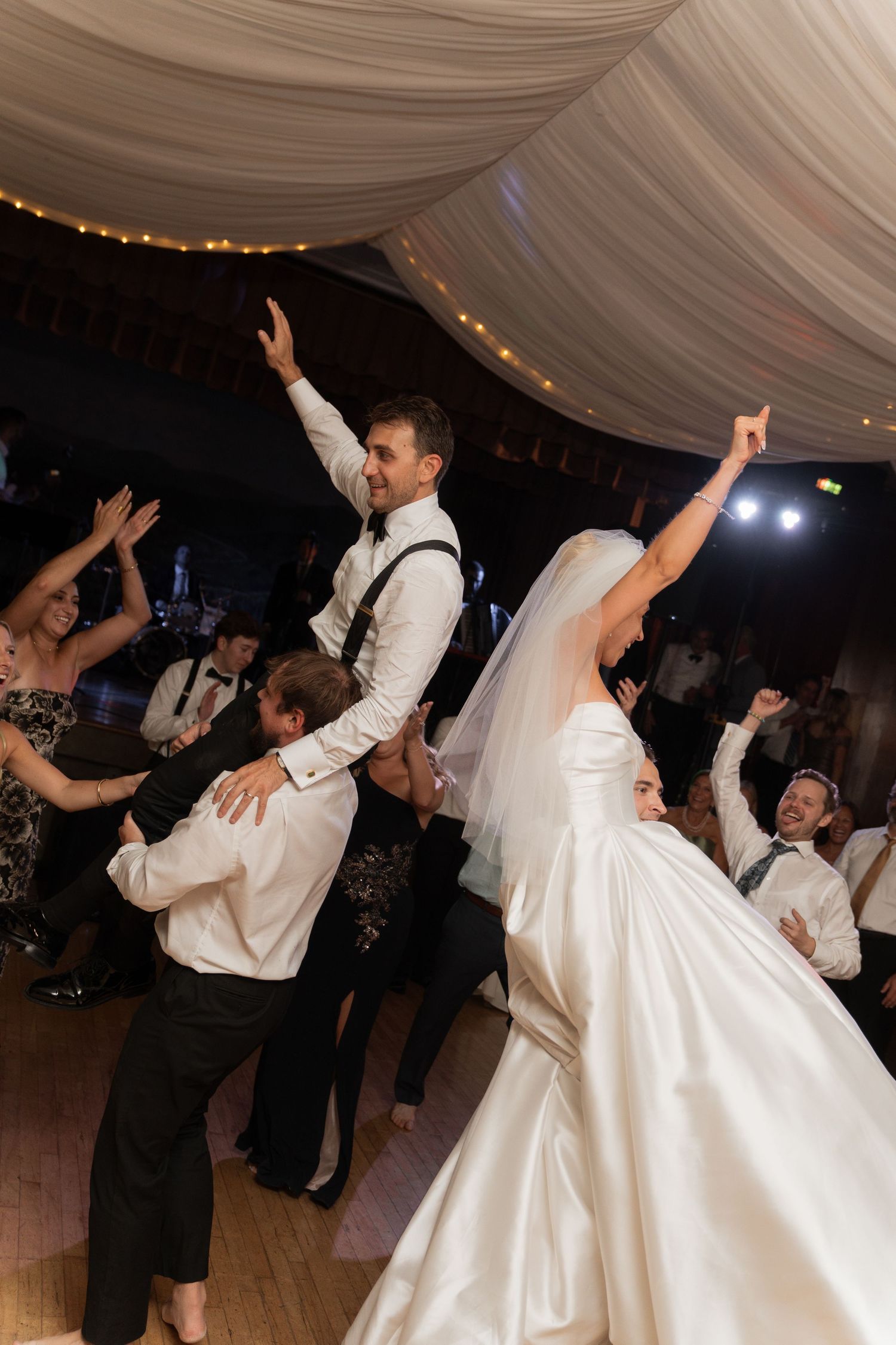 Bride and groom joyfully dancing with raised arms at their wedding reception surrounded by guests.
