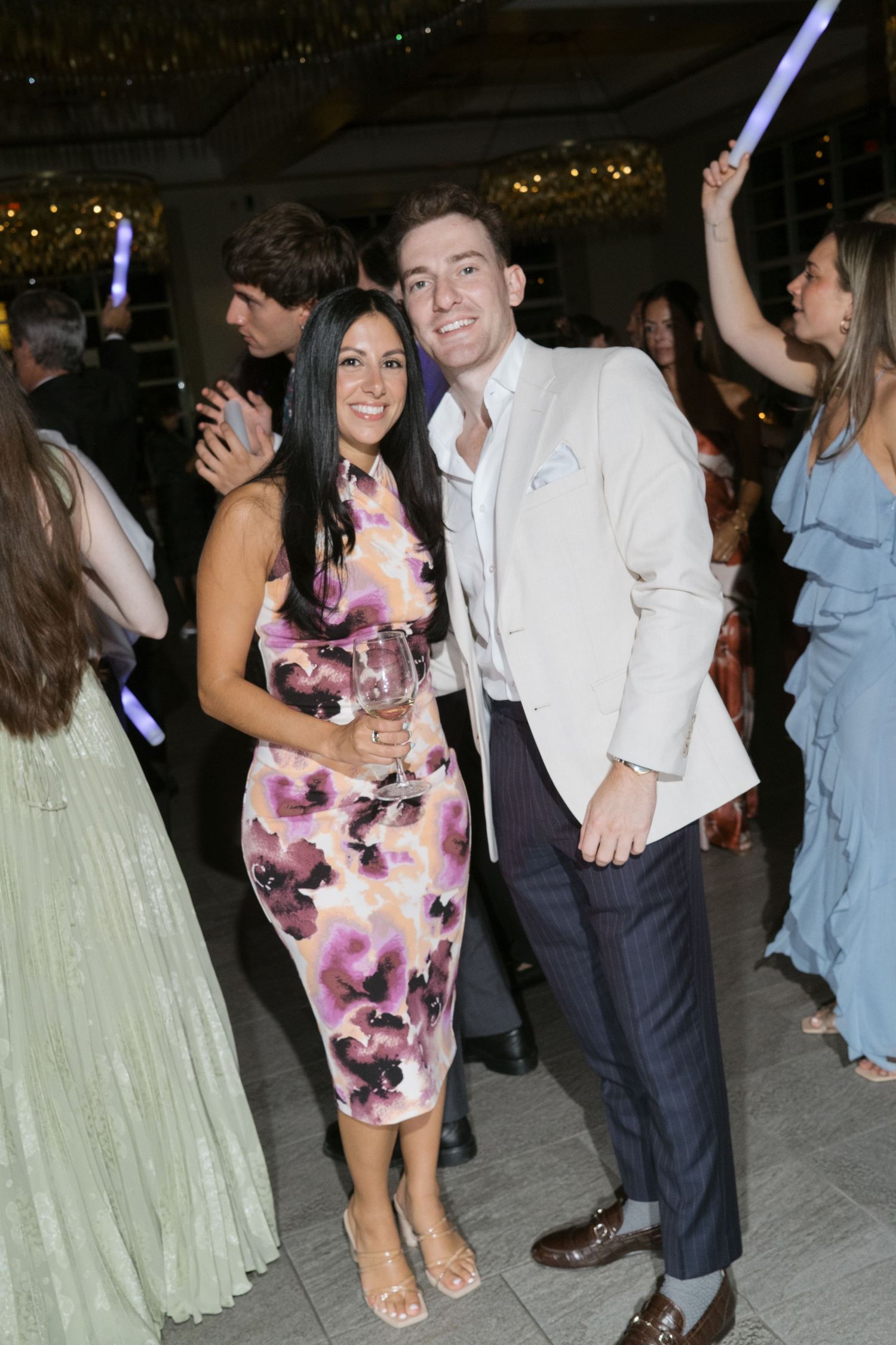 Couple posing together at a wedding reception, woman in floral dress and man in white blazer on the dance floor.