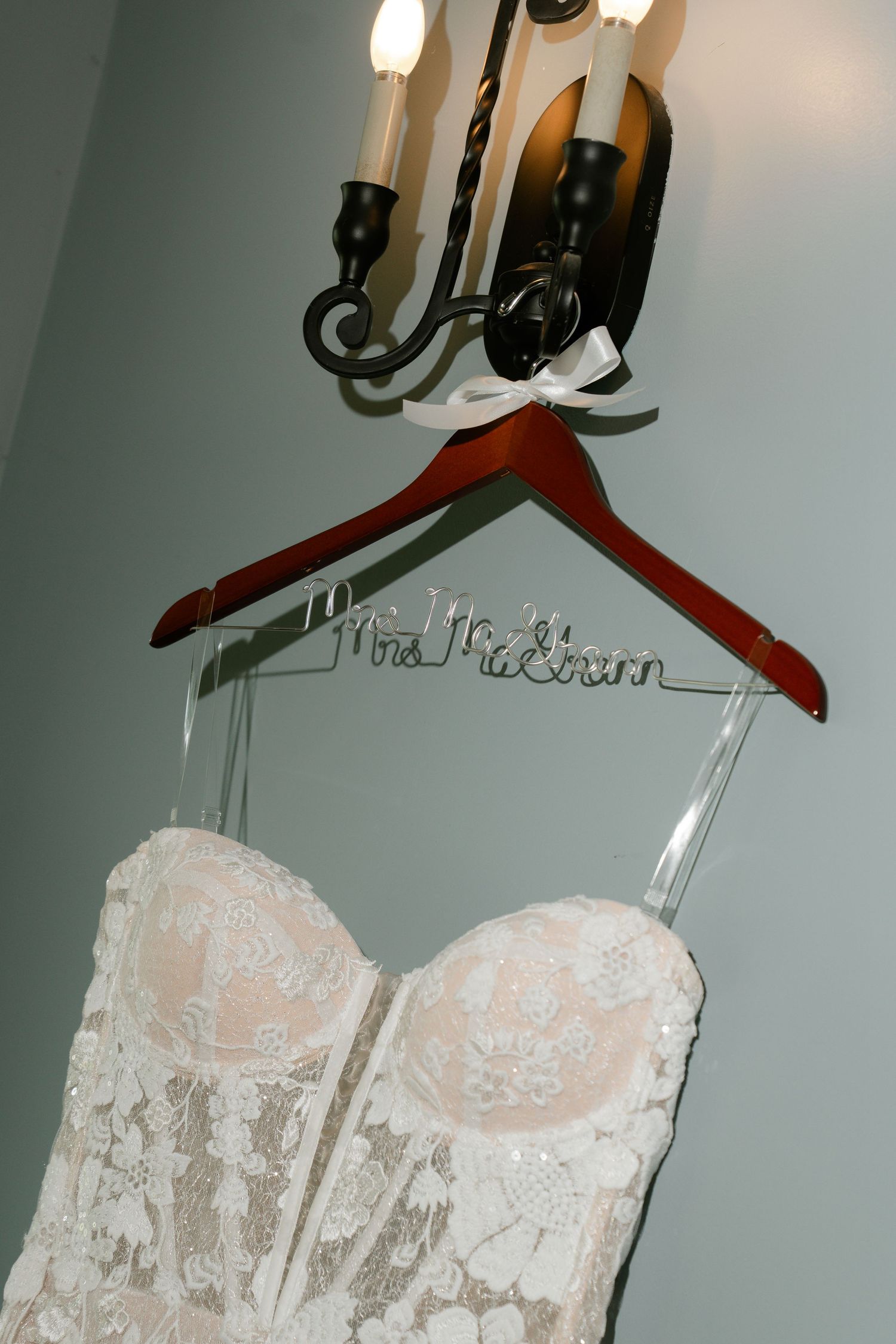 Elegant white lace wedding dress with sweetheart neckline hanging on wooden hanger against grey wall.
