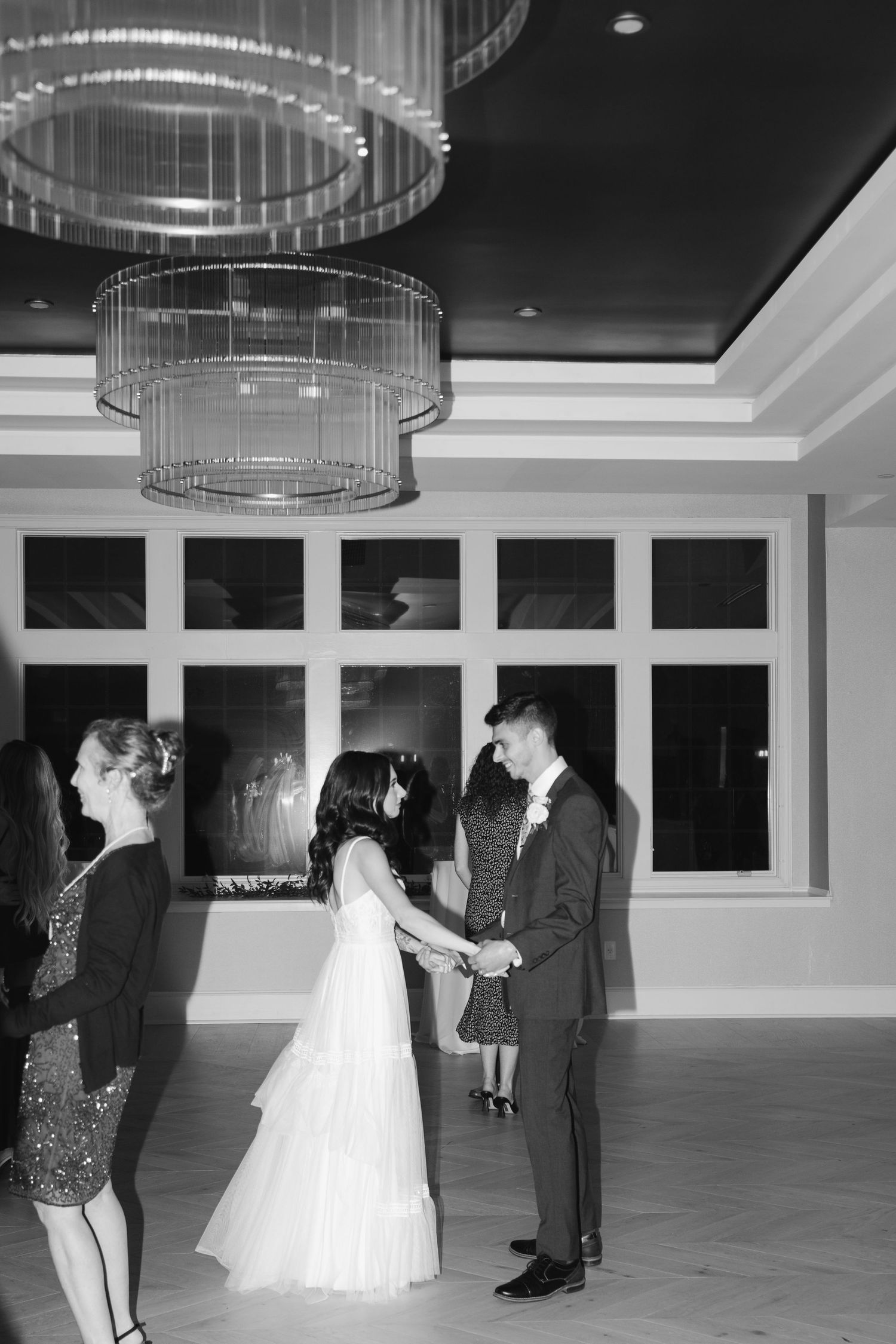 A black and white photo of a wedding couple's first dance under elegant crystal chandeliers in a modern ballroom.
