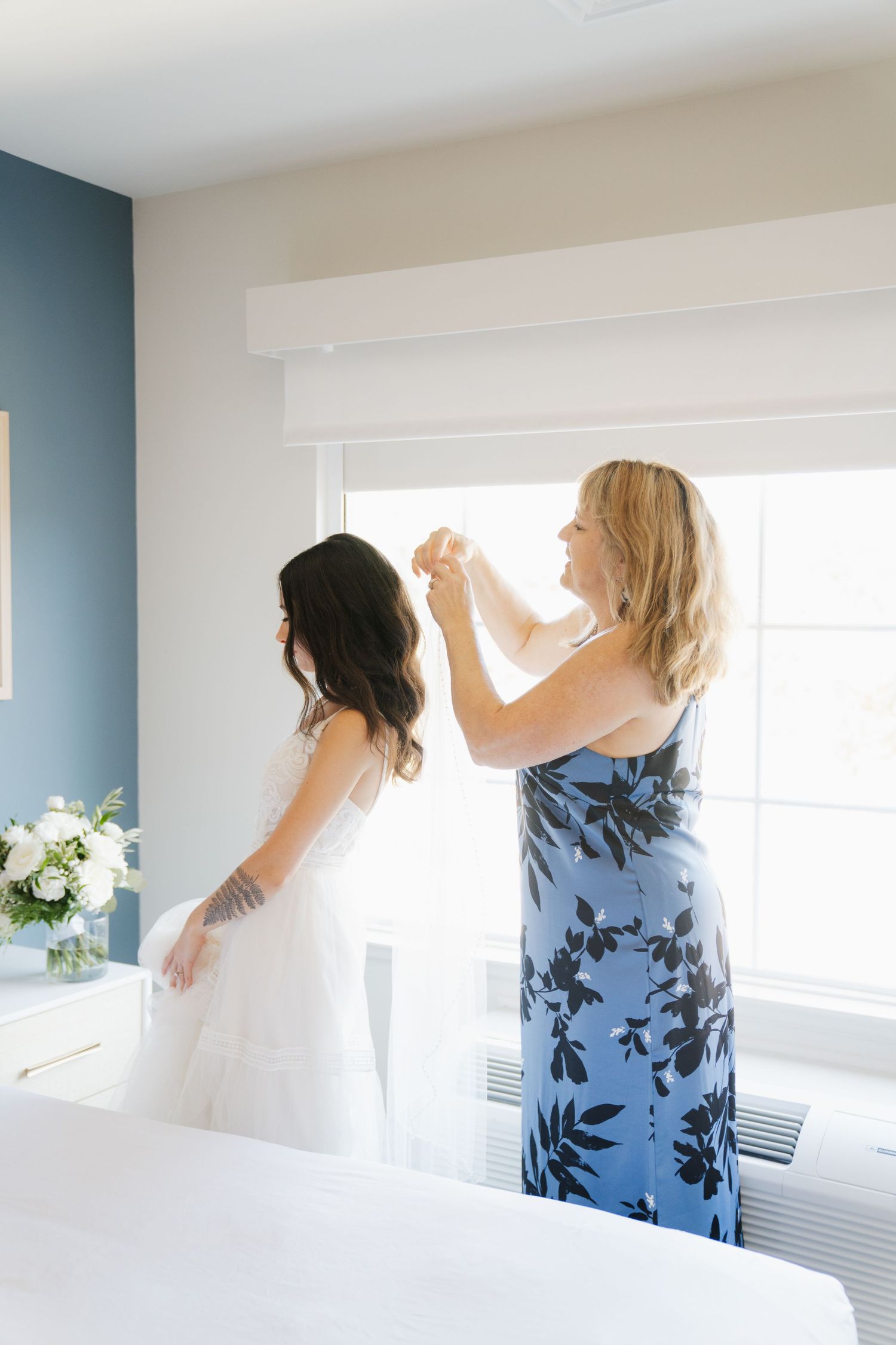 A woman in a blue floral dress helps adjust a white wedding dress by a bright window.