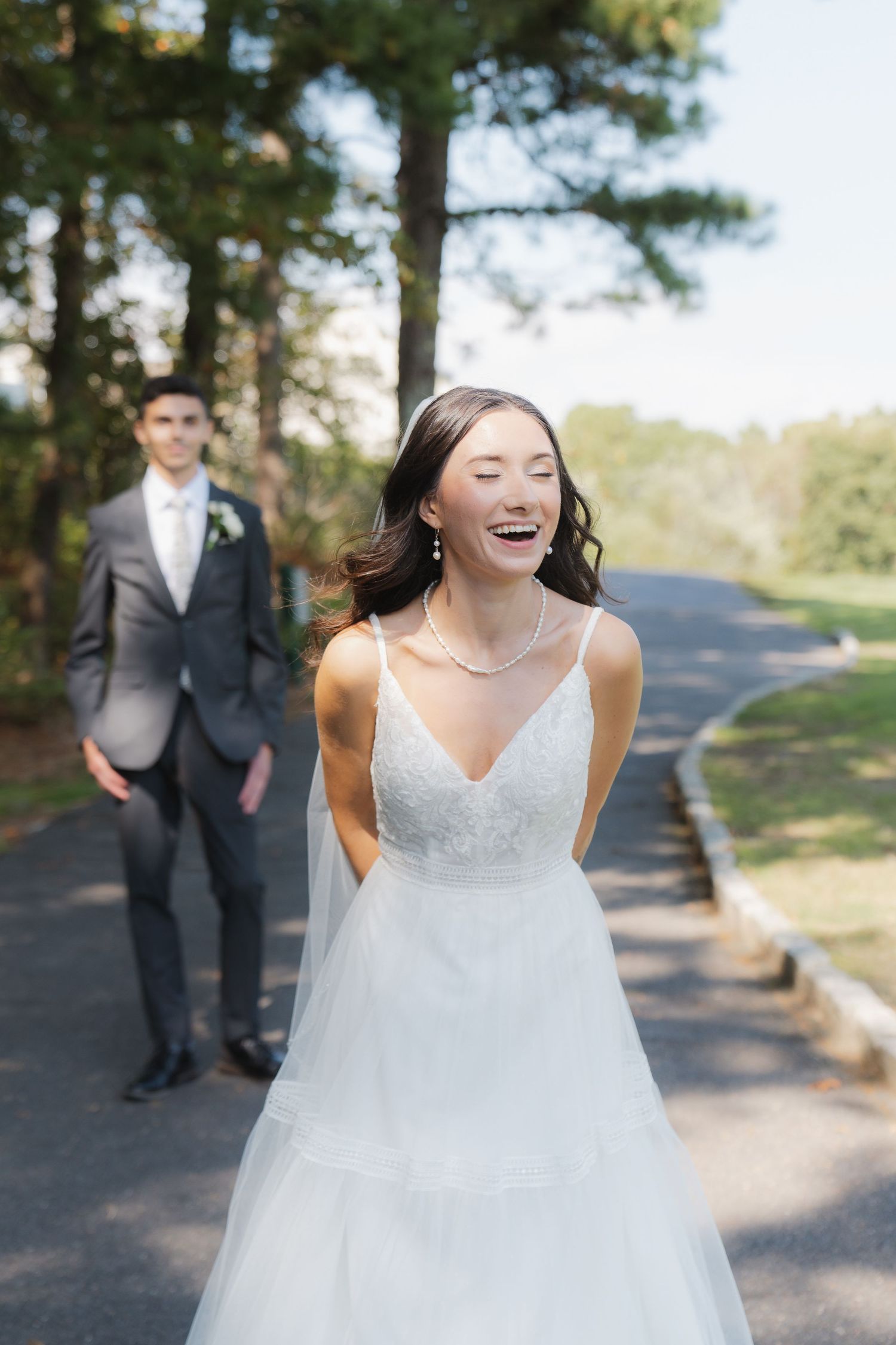 A joyful bride in a flowing white dress laughs on a tree-lined path while a groom stands in the background.