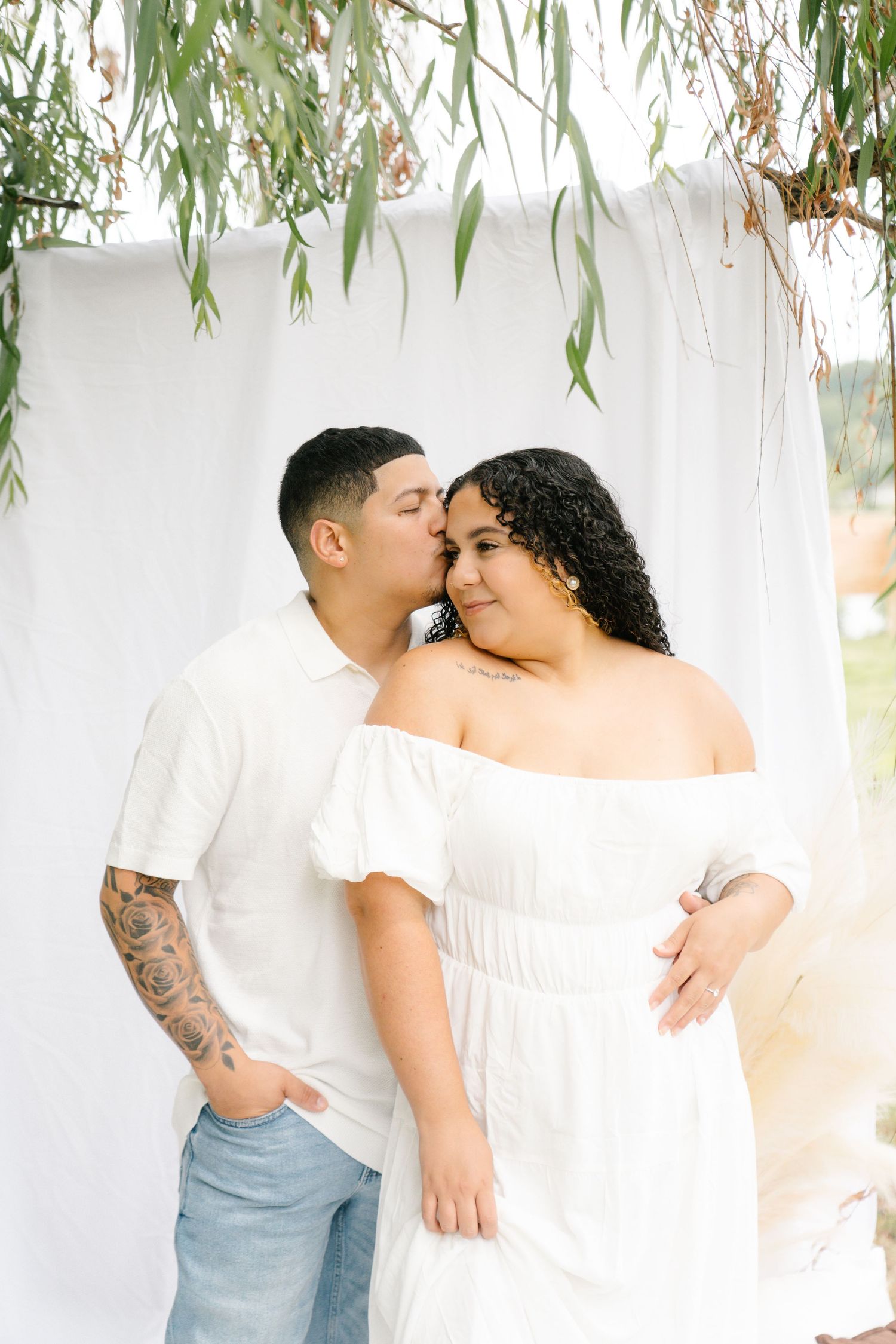 A romantic couple sharing tender moments under hanging willow branches with a white backdrop.