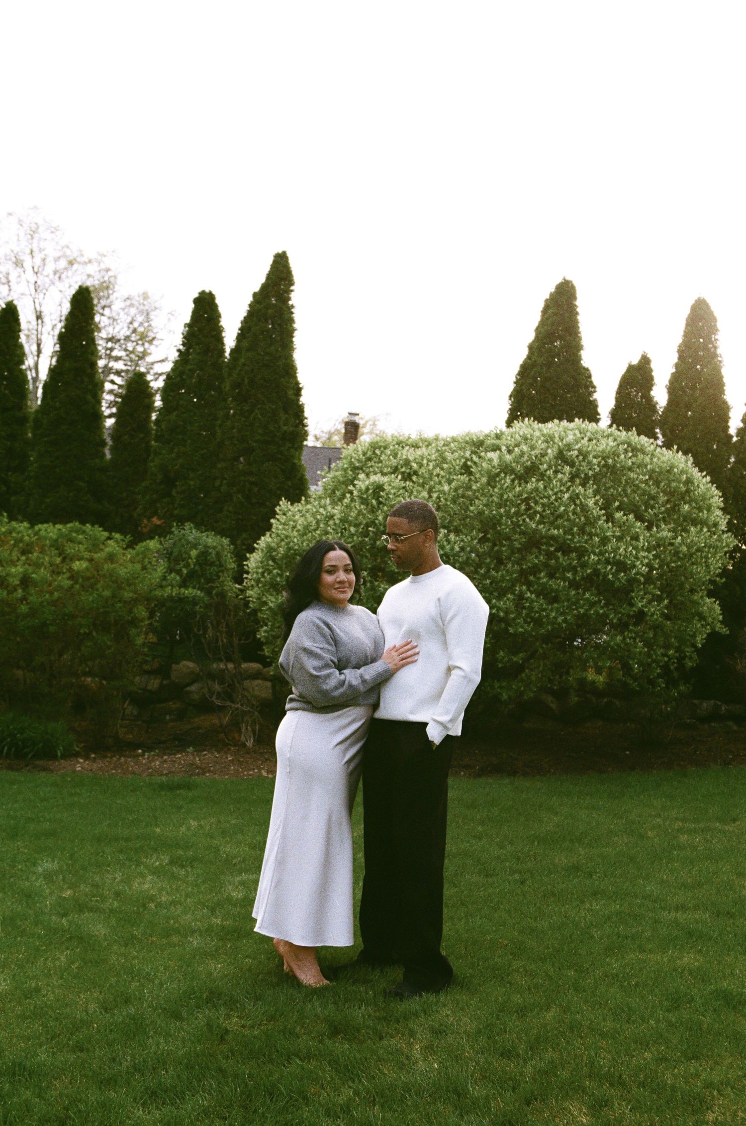 Couple posing in lush green garden, woman in gray dress with baby bump, man in white shirt and black pants.