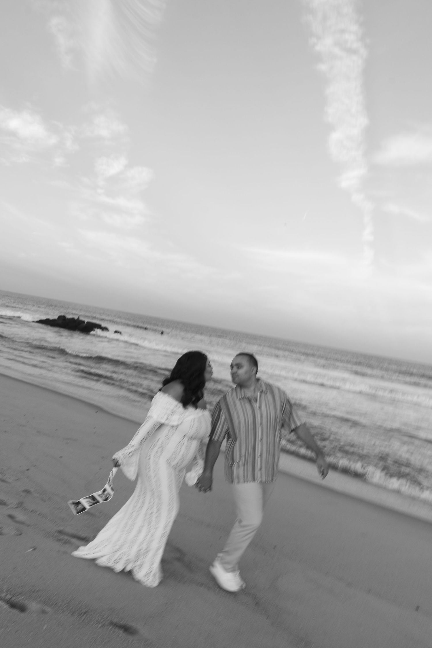 Black and white photo of a couple walking hand in hand on a beach at sunset, woman in white dress.