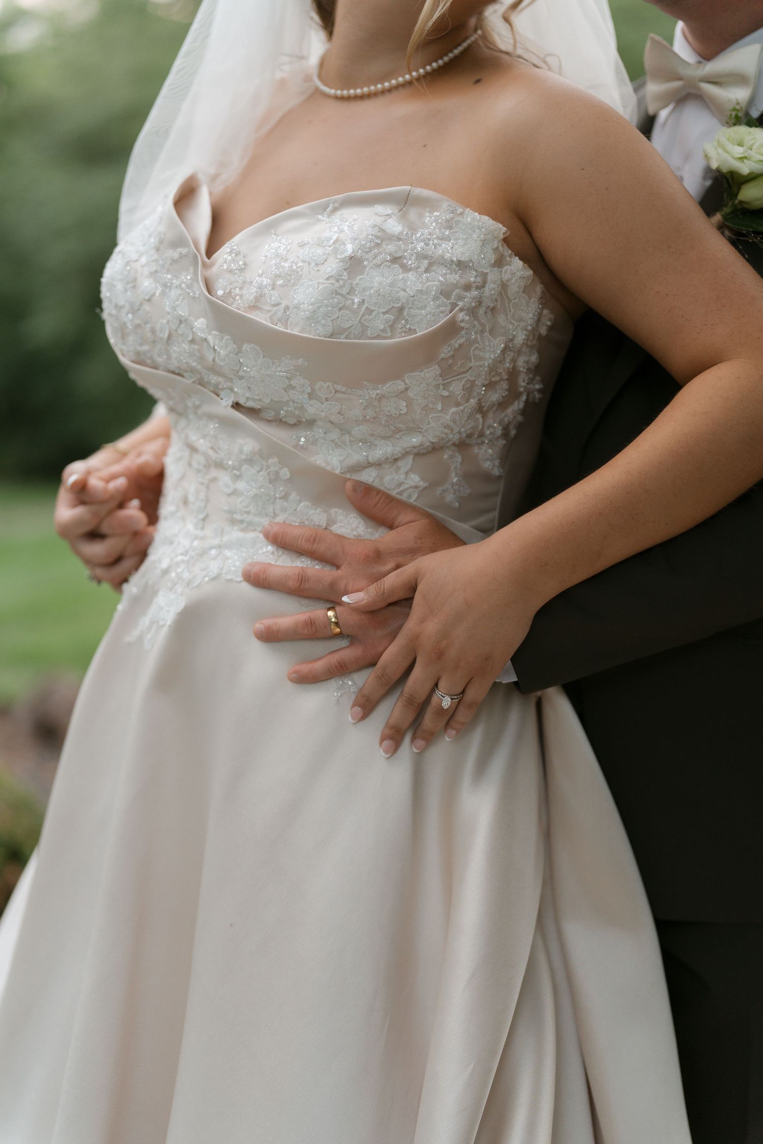Close-up of bride in strapless lace wedding gown with groom embracing her waist, showing wedding rings.