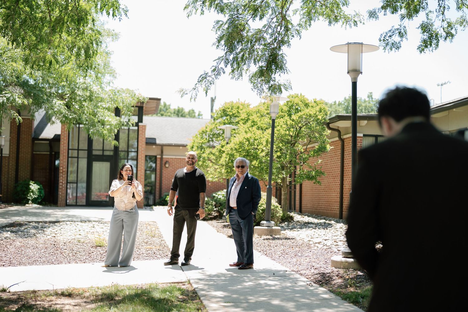 Three people stand on a sunny outdoor campus courtyard path surrounded by trees and a modern building.