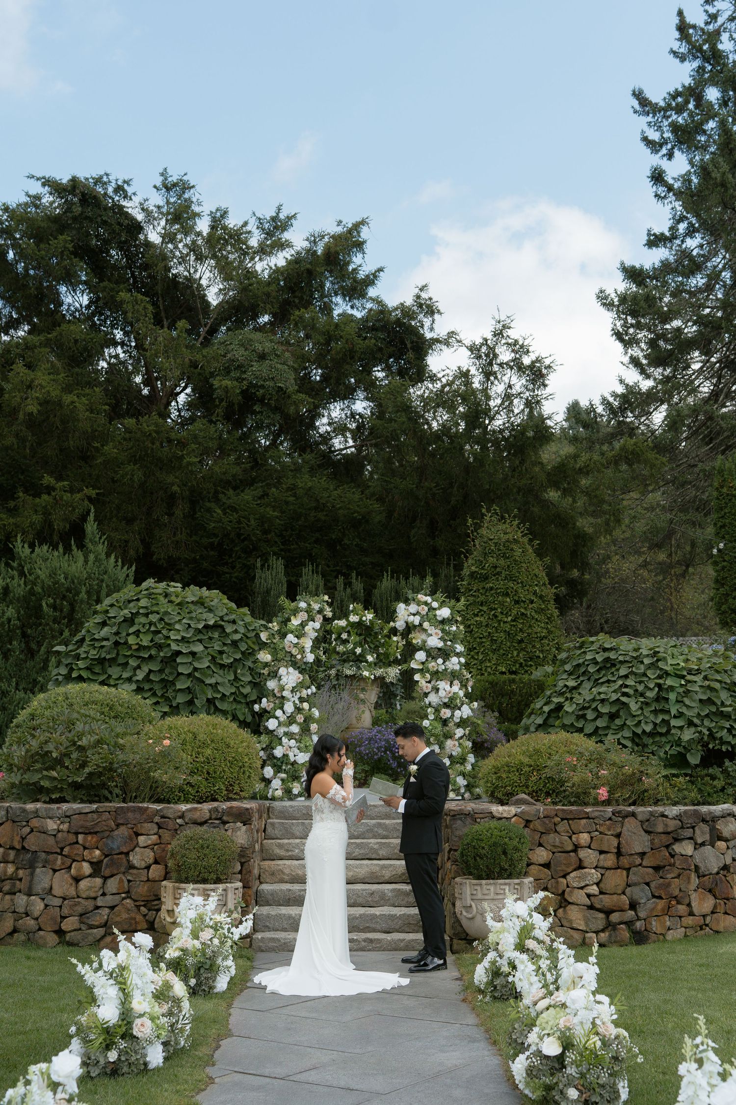 Bride and groom exchange vows under floral arch with white flowers at elegant outdoor garden wedding ceremony.