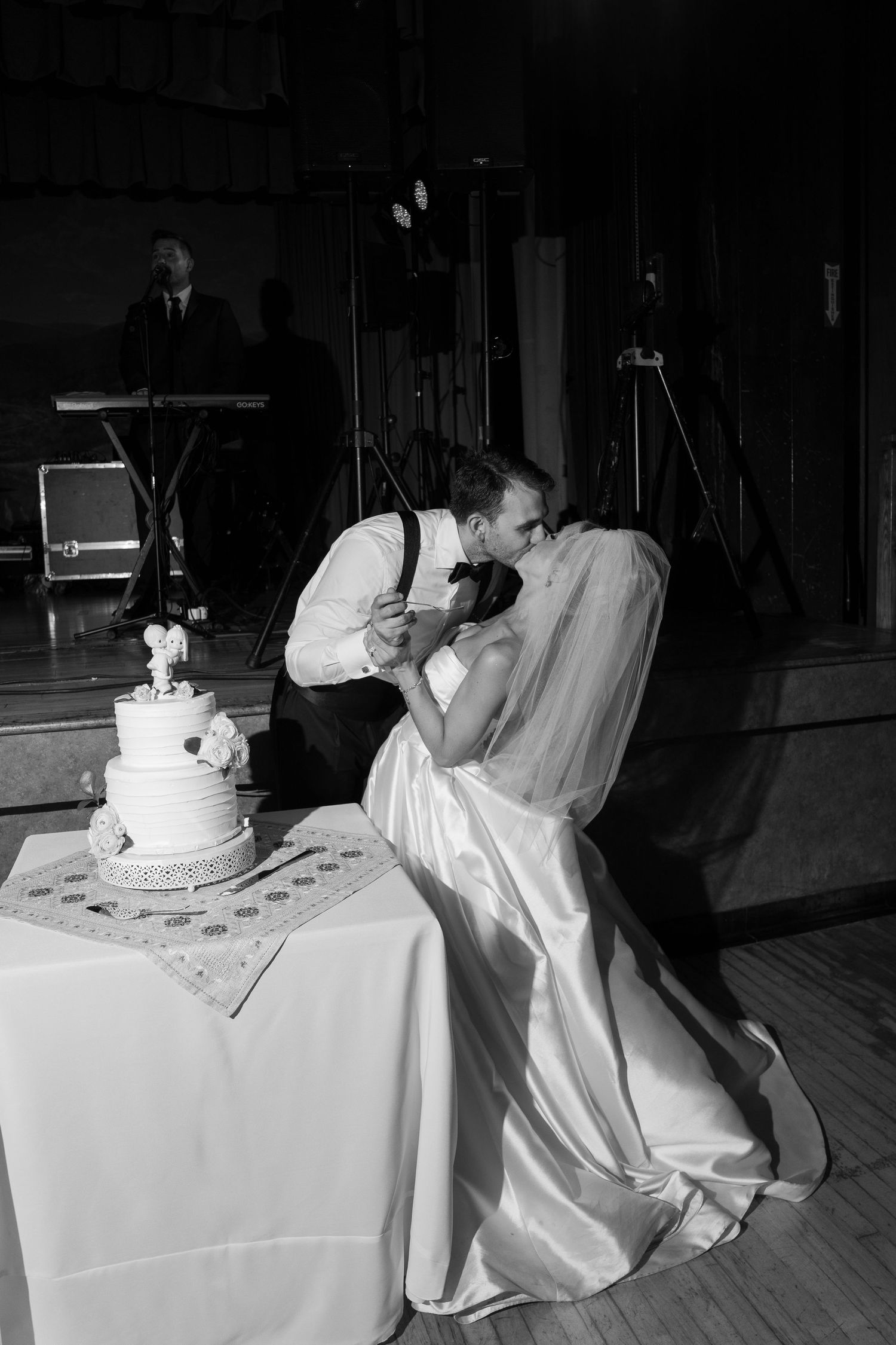 Black and white photo of bride and groom sharing a kiss beside tiered wedding cake at reception.