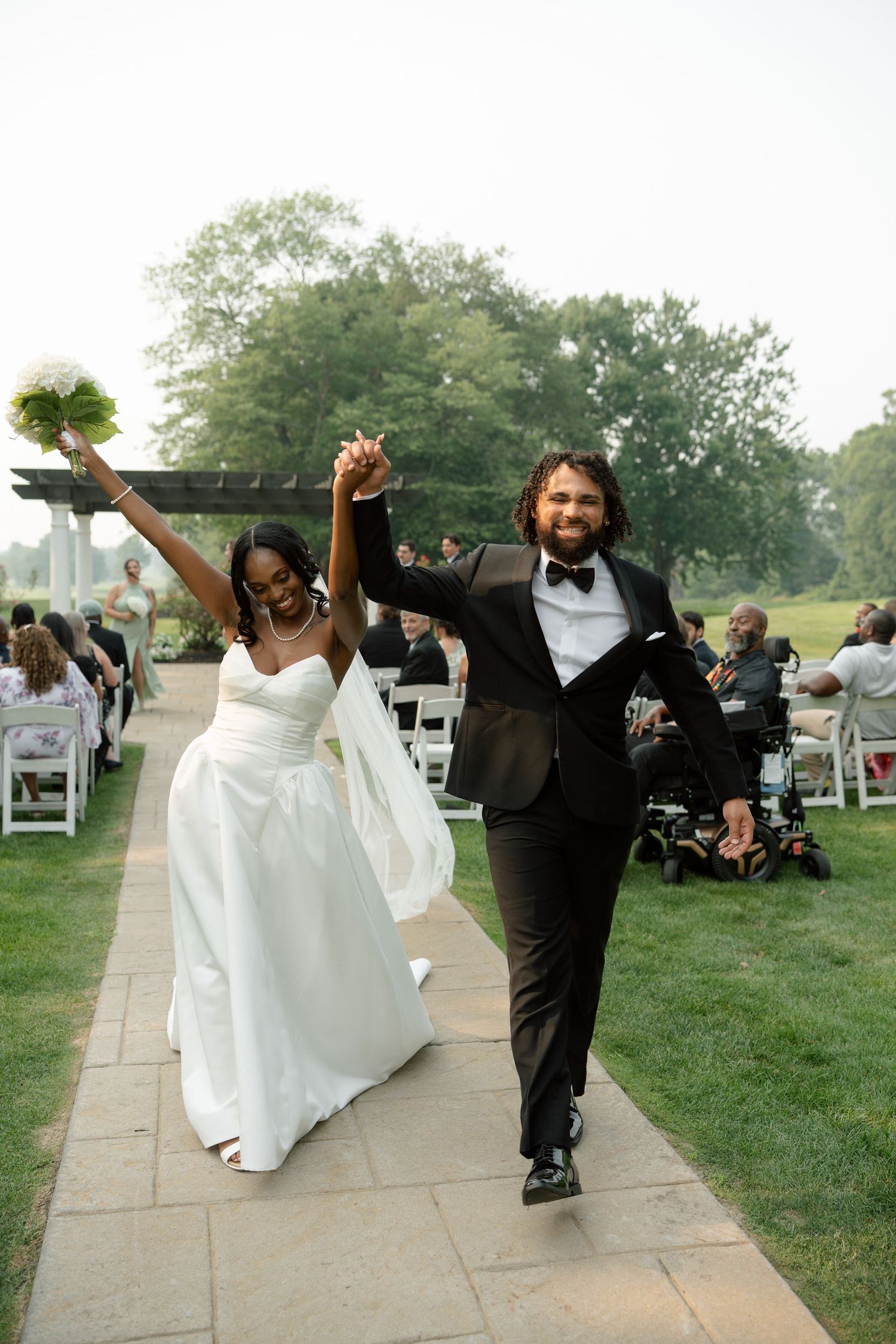 Newlyweds celebrate recessional holding hands with bride raising bouquet triumphantly at outdoor ceremony.
