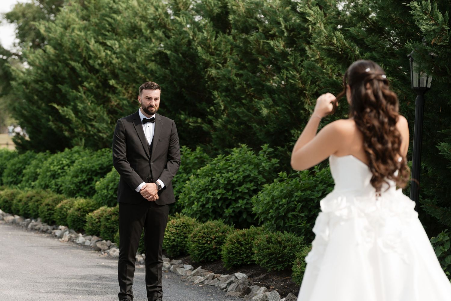 Groom in black tuxedo sees bride in white gown for first look at outdoor wedding venue.