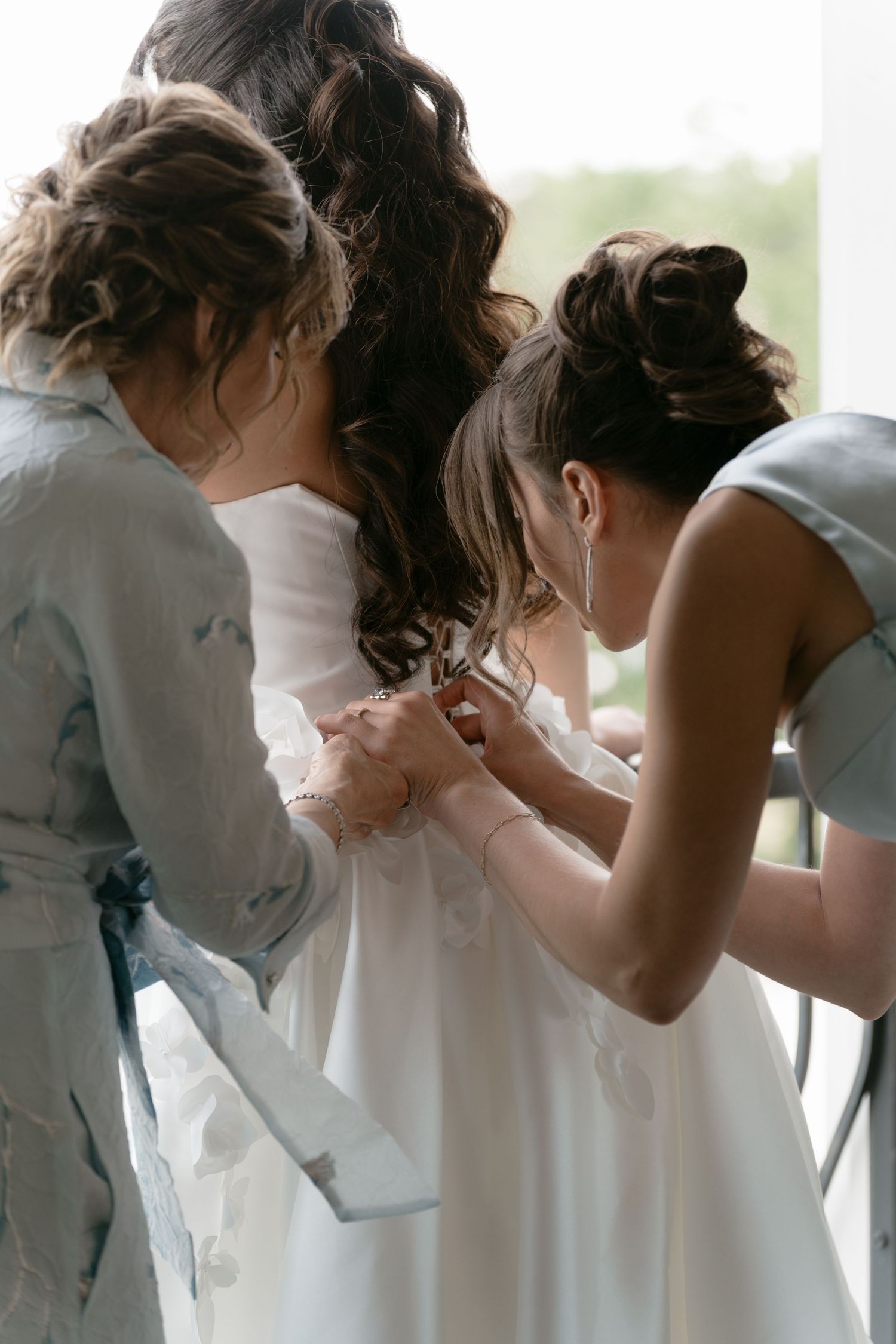 Two bridesmaids in sage gowns help fasten the brides white wedding dress from behind.