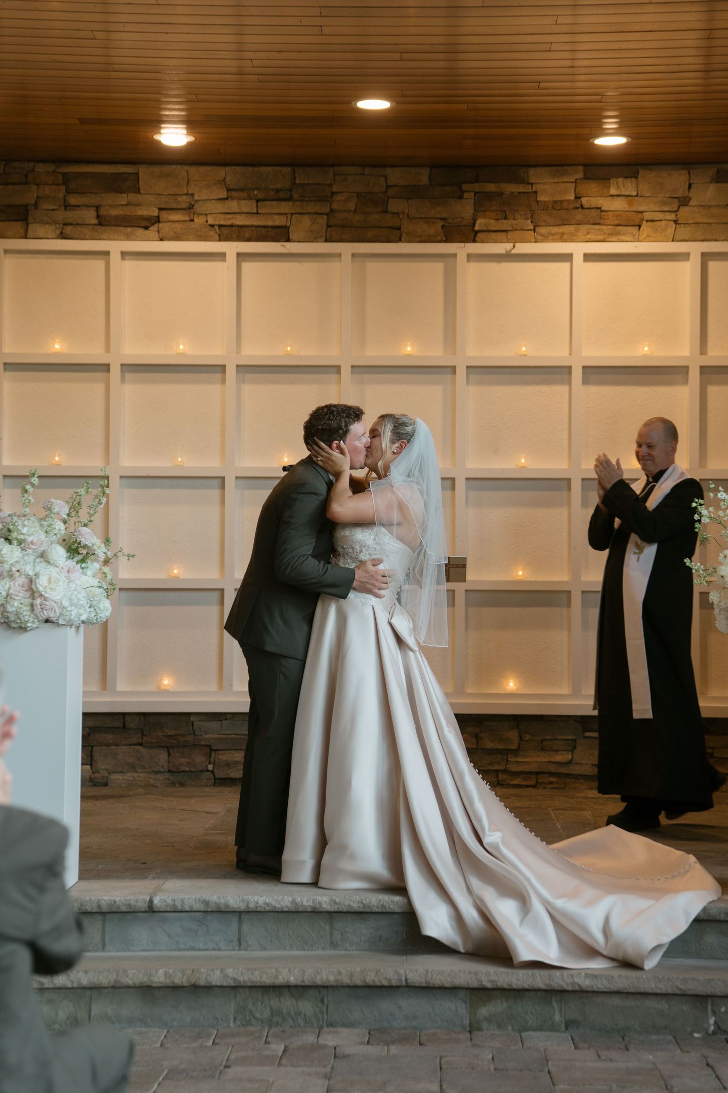 Bride and groom share first kiss at elegant wedding ceremony with candlelit white wall backdrop and officiant.