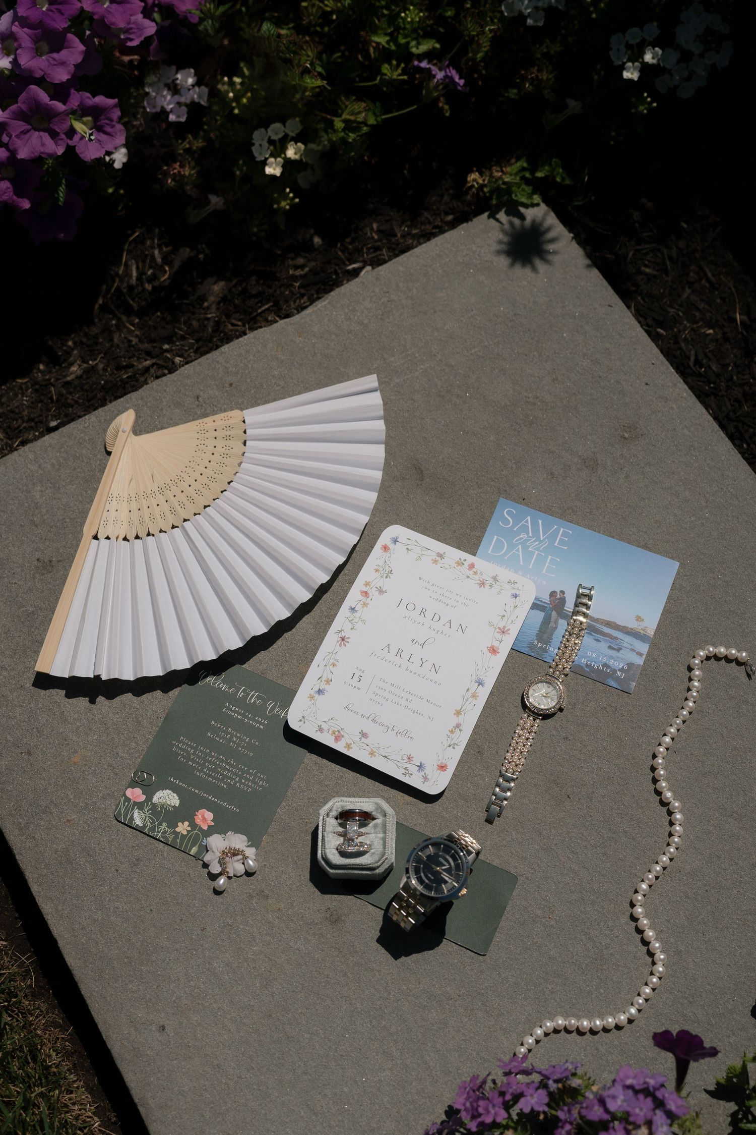Elegant wedding flat lay featuring floral invitations, pearl necklace, watches, rings, and a white hand fan on gray stone.