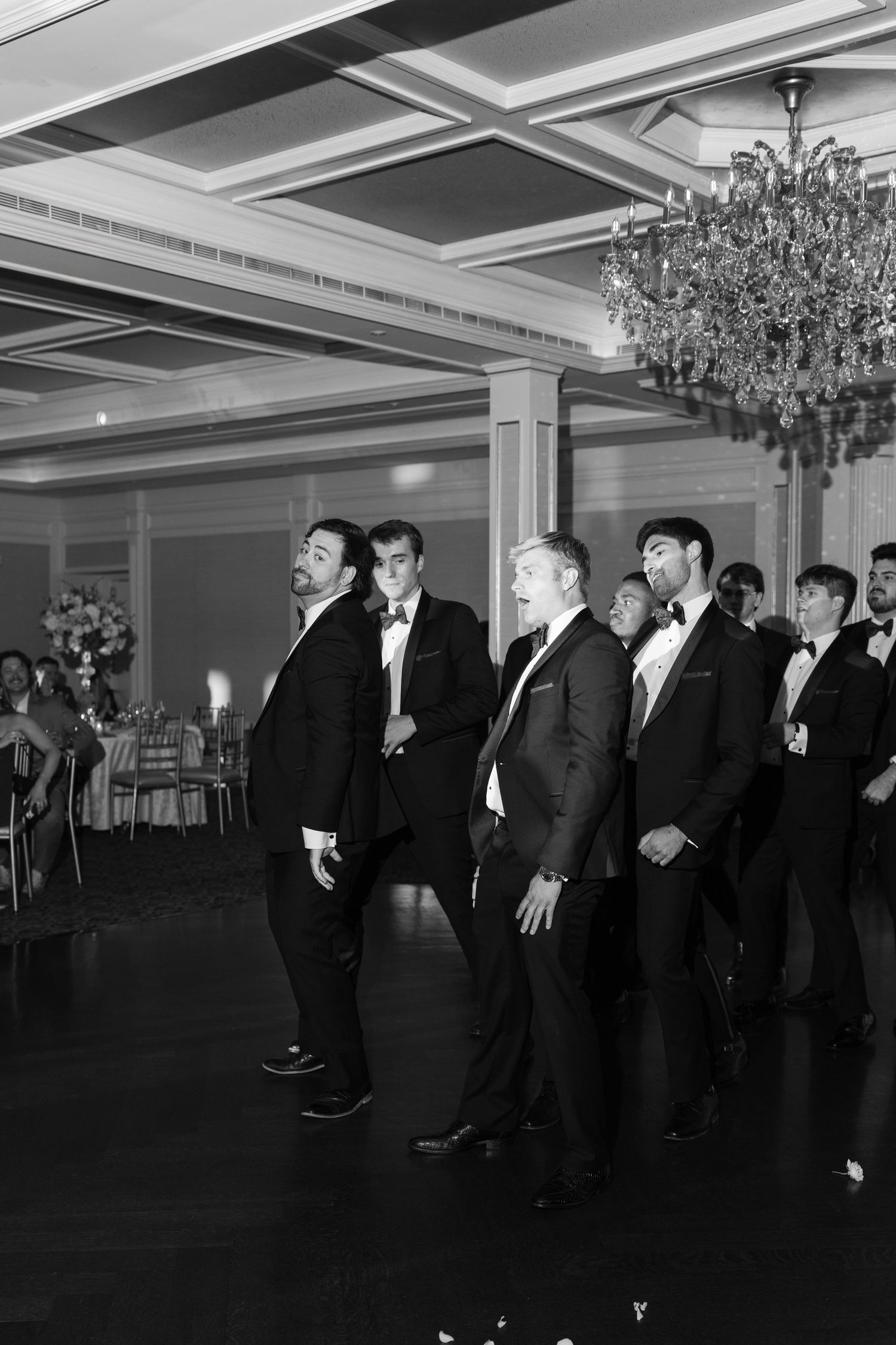 Groomsmen in black tuxedos performing a choreographed dance routine in elegant ballroom at wedding reception.