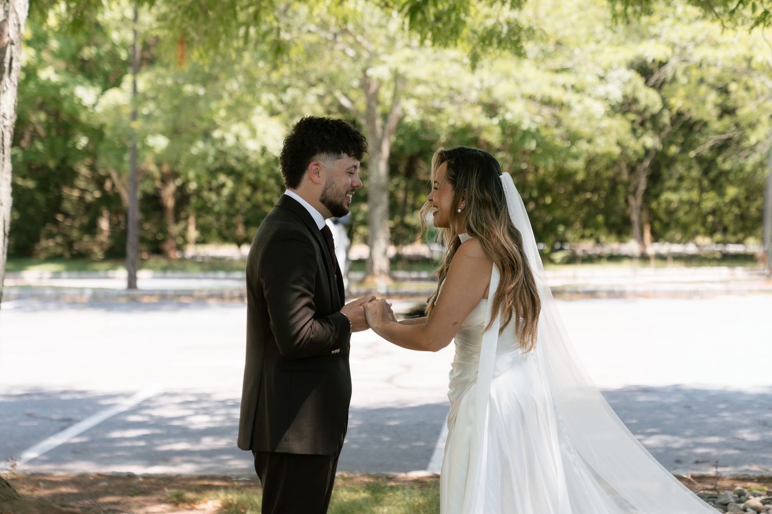 A bride and groom hold hands outdoors in a park during their wedding ceremony, surrounded by lush green trees.