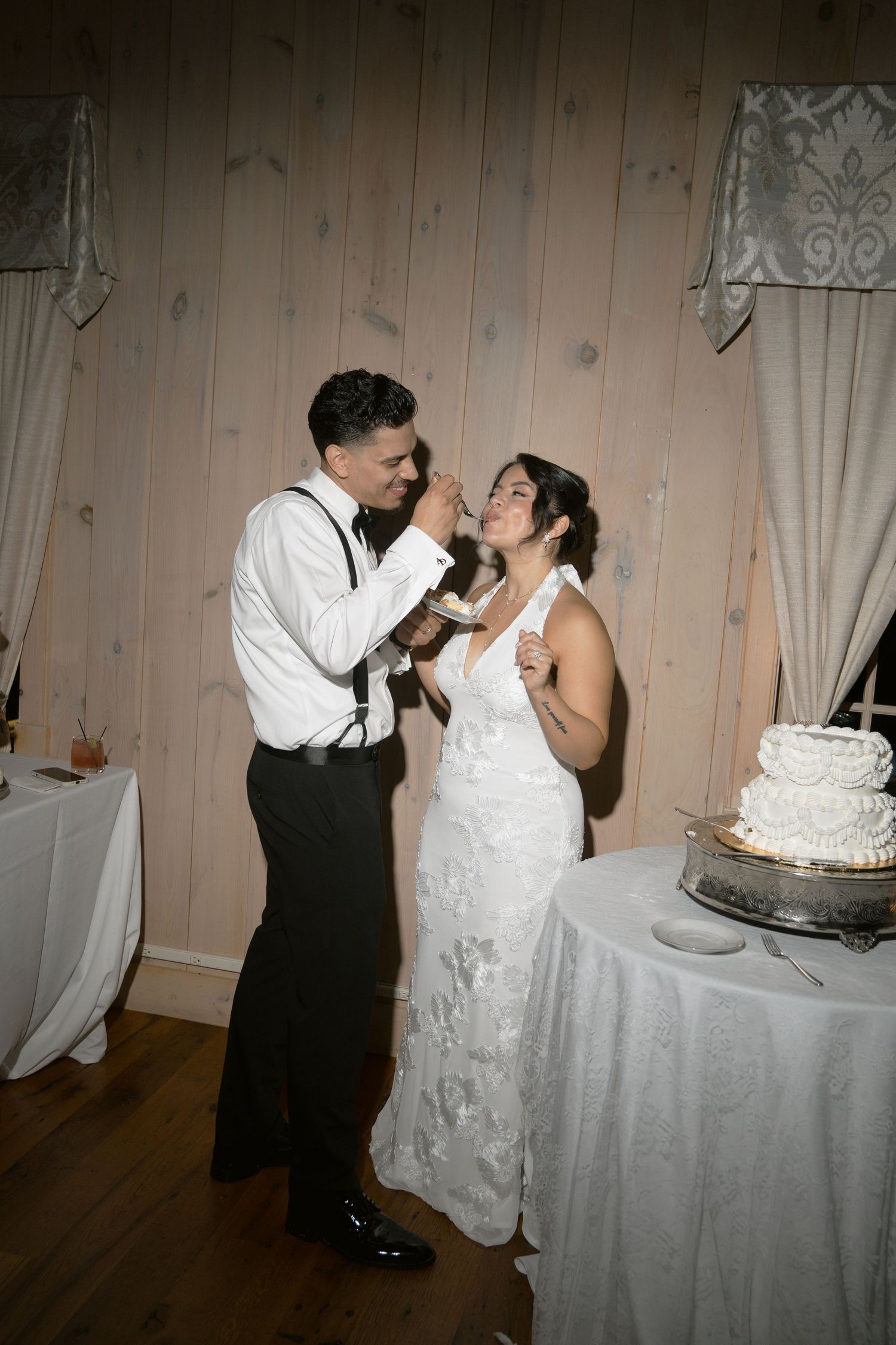 Bride and groom laugh during wedding cake cutting at rustic indoor reception venue with white tiered cake.