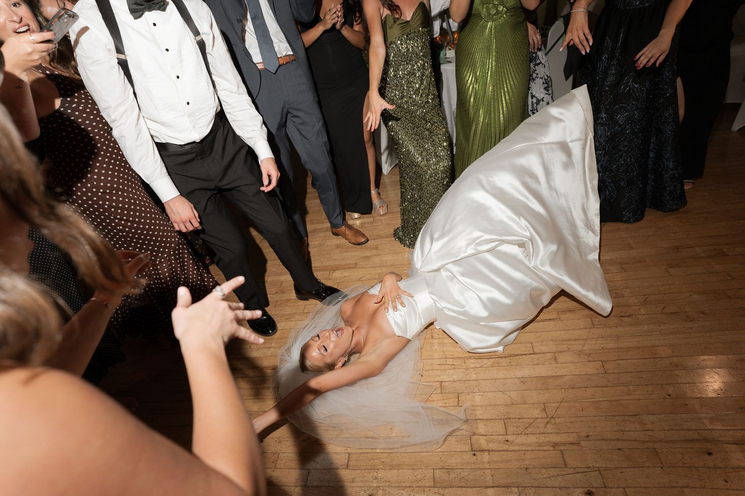 Bride in white satin gown dancing on wooden floor at lively wedding reception celebration.