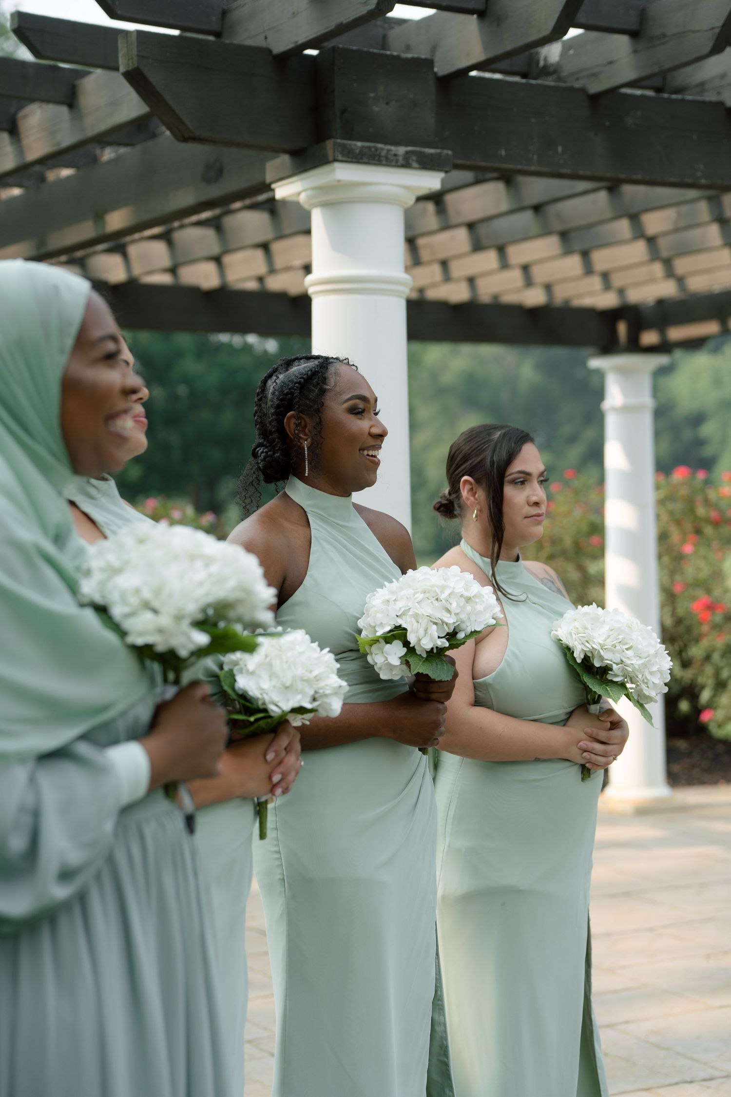 Four bridesmaids in sage green dresses holding white hydrangea bouquets stand under a pergola at an outdoor wedding.