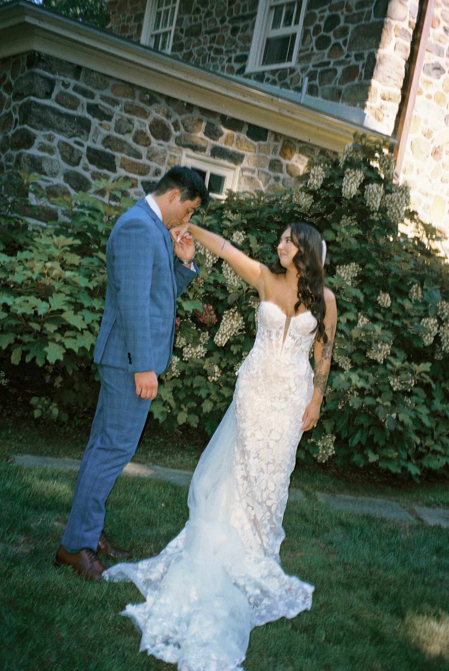 A couple poses for wedding photos in front of a stone building with lush greenery.