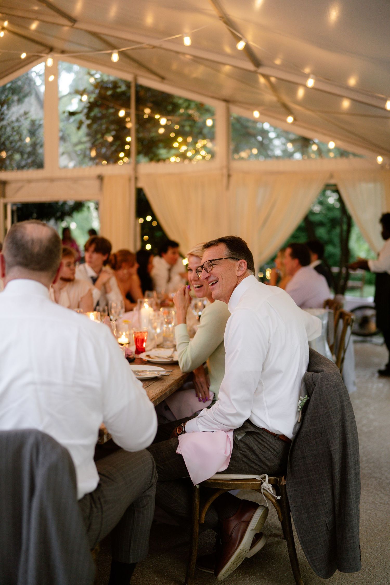 Wedding reception guests enjoying dinner under string lights in an elegant white tent setting.