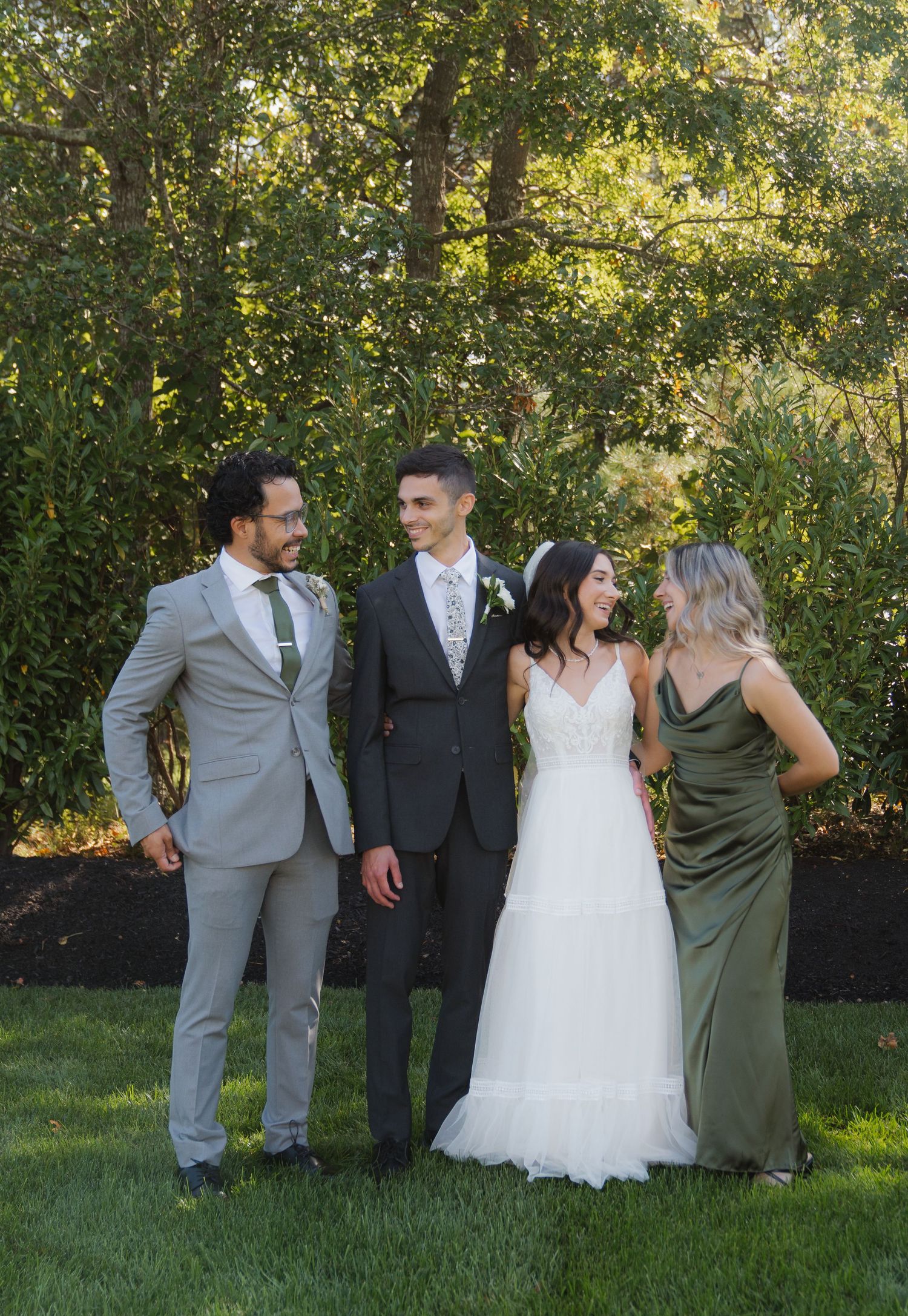 Wedding party members in gray and green attire pose together on a sunny lawn with trees in the background.