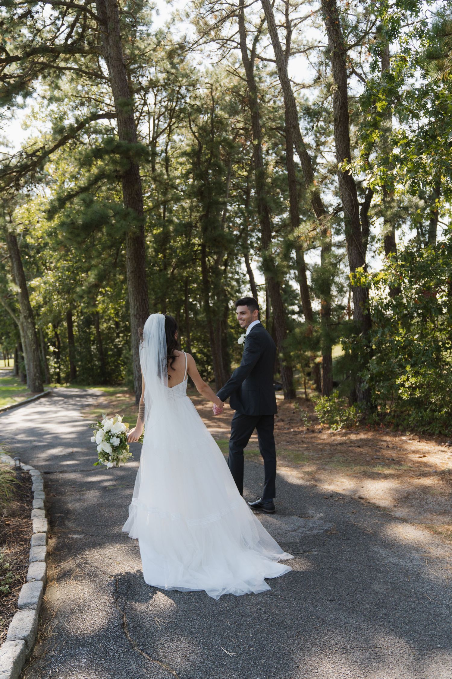 A couple in wedding attire share a romantic moment on a tree-lined gravel path during their outdoor photo session.