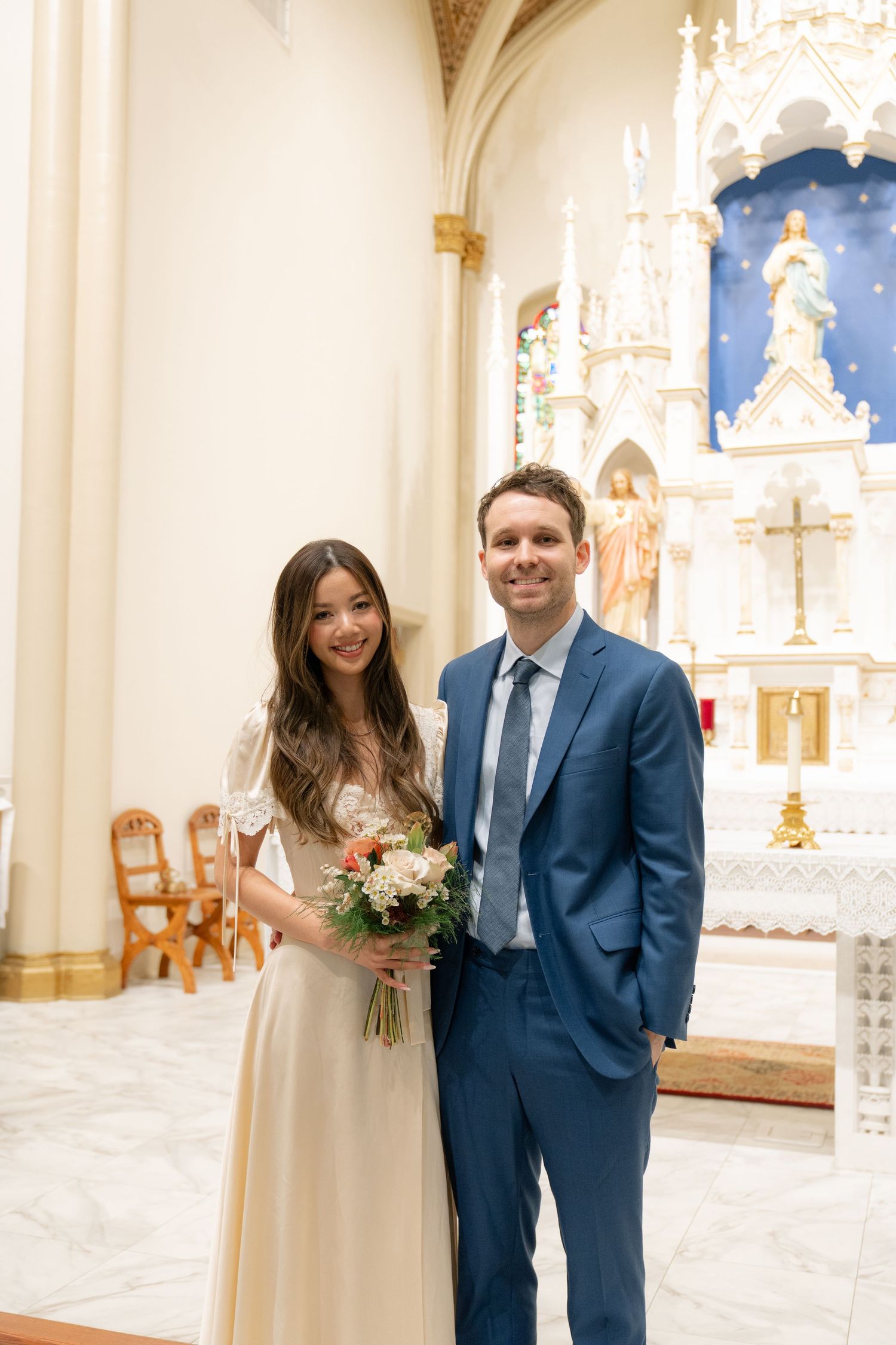 A couple poses for a wedding photo inside an ornate church with white walls and religious decor.
