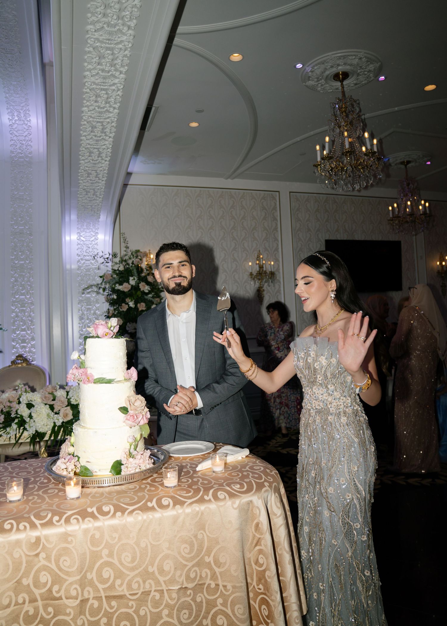 Wedding celebration with guests gathered around a white tiered cake on an ornate tablecloth.