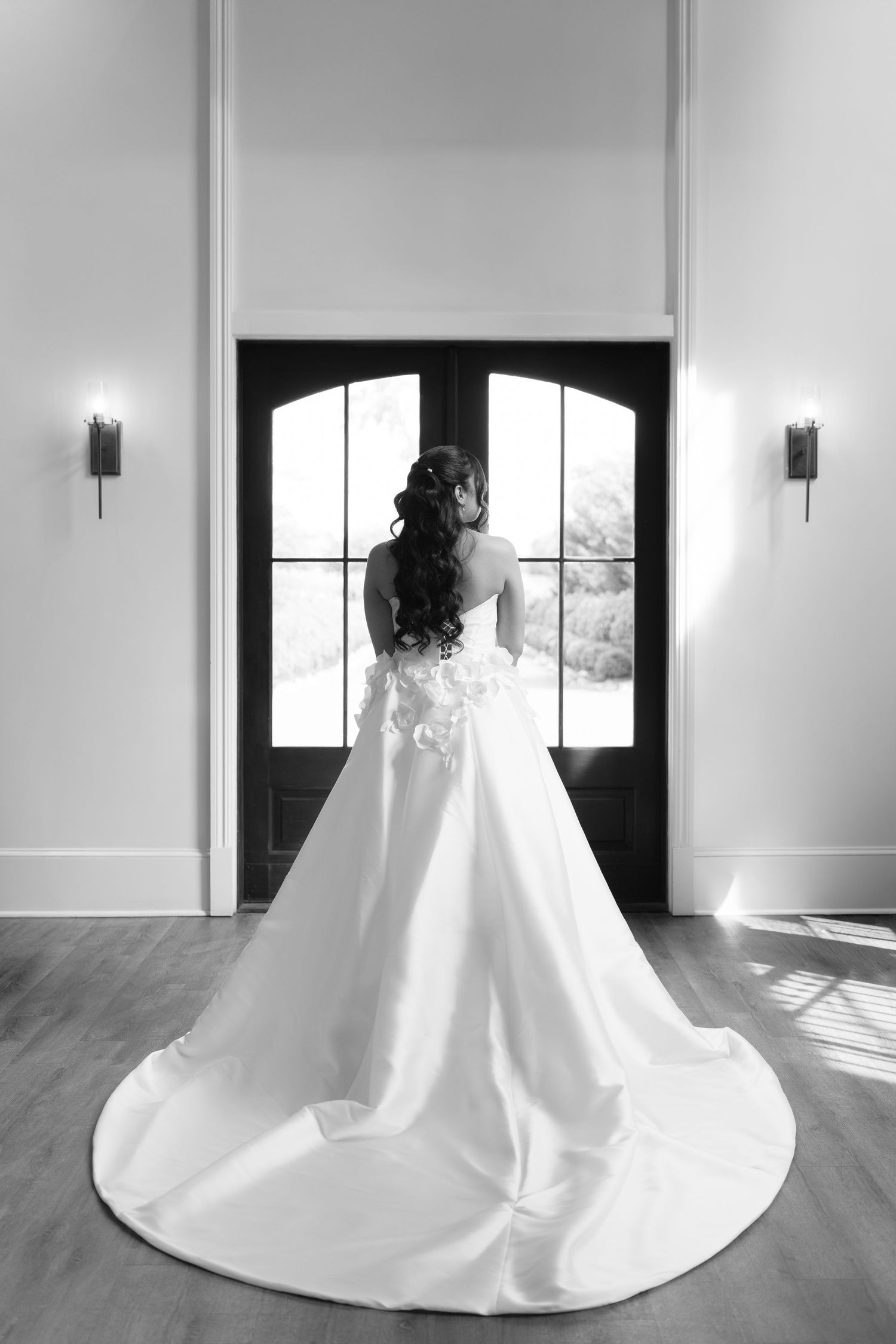Black and white photo of bride in ball gown with long train standing before double doors.