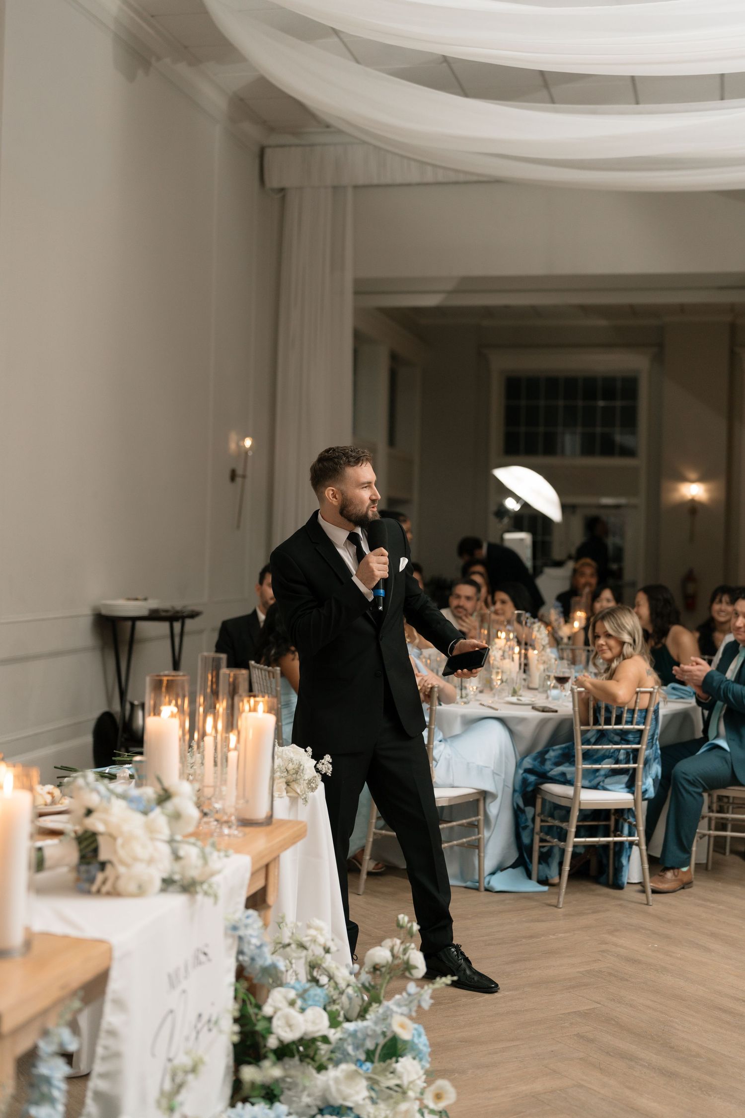 Groom in black tuxedo gives speech at wedding reception beside floral arrangements and candlelit tables.