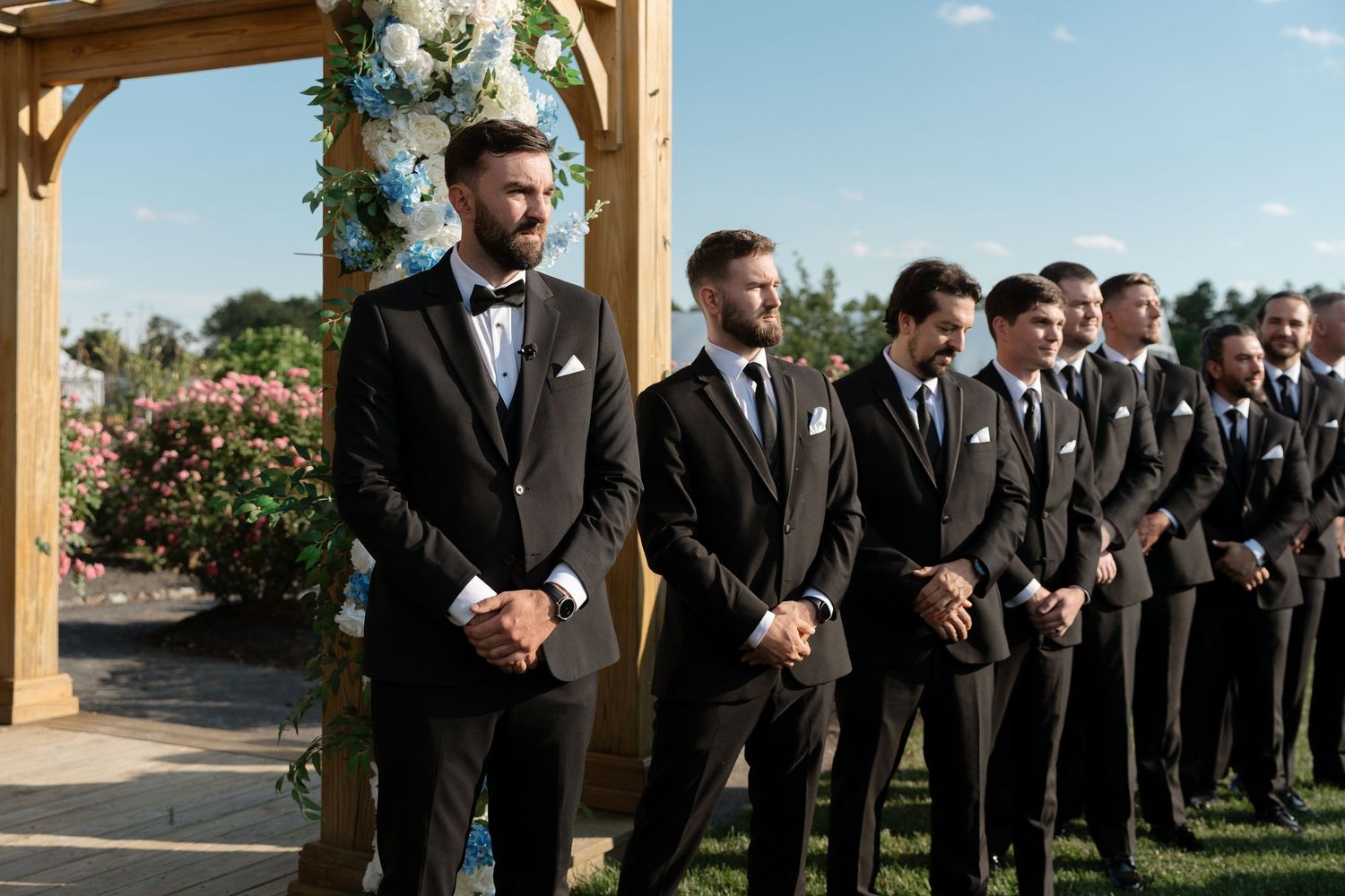 Groom and groomsmen in black tuxedos standing at floral wooden arch during outdoor wedding ceremony.