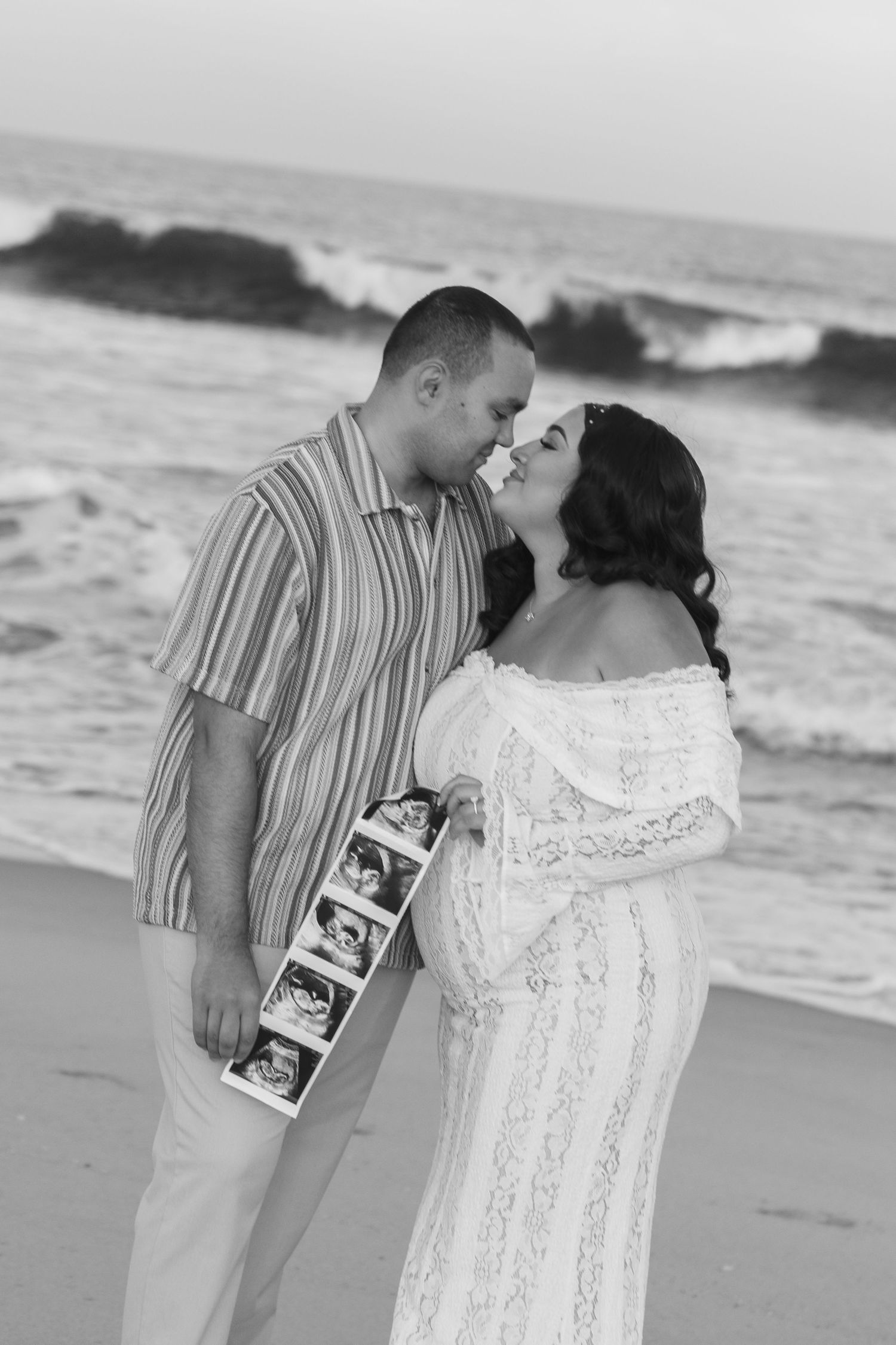 Pregnant couple sharing a romantic moment on the beach holding ultrasound photos in black and white.
