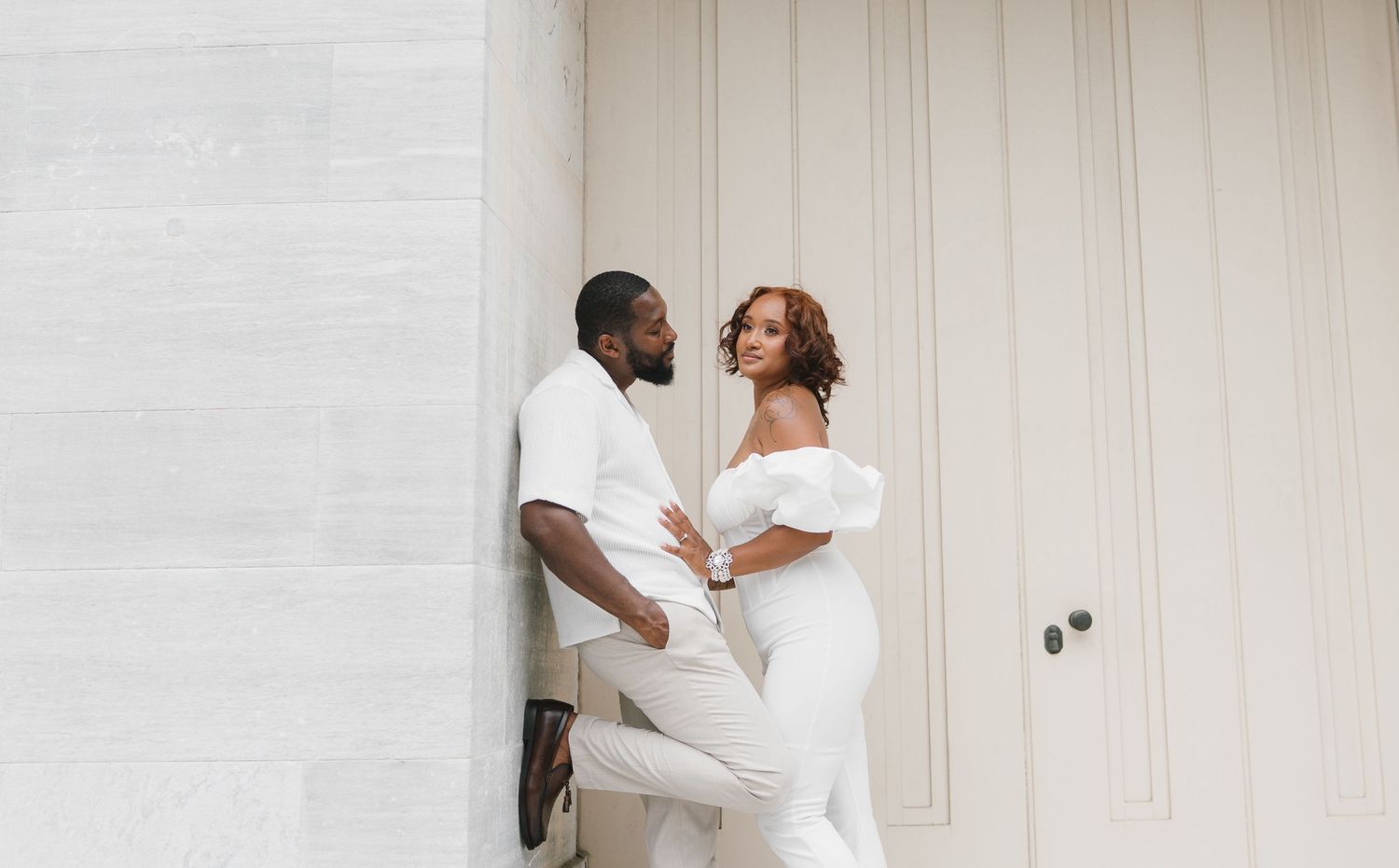 A couple in matching white outfits share an intimate moment against a light-colored wall.