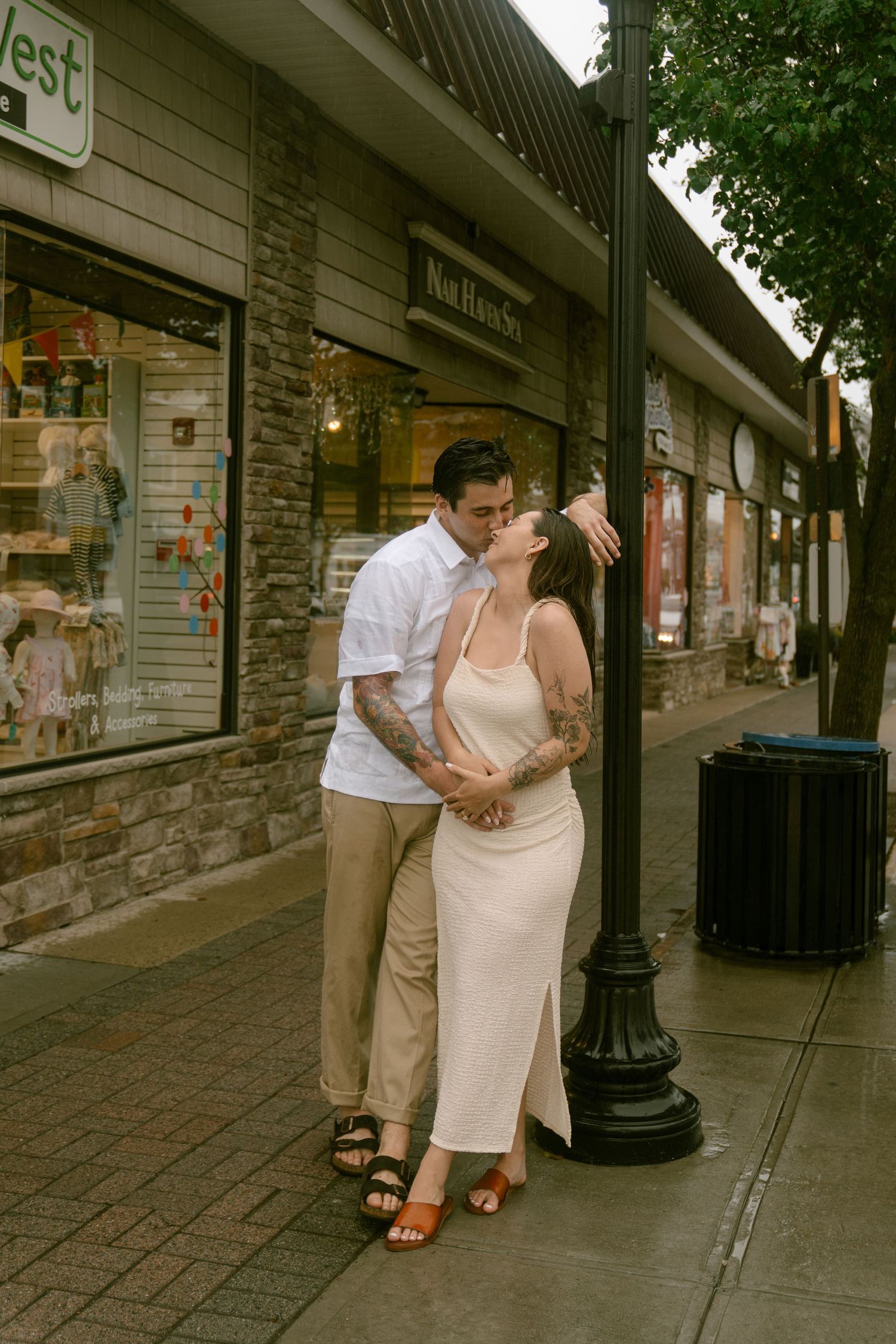A romantic couple embraces on a sidewalk outside storefronts in casual summer attire.