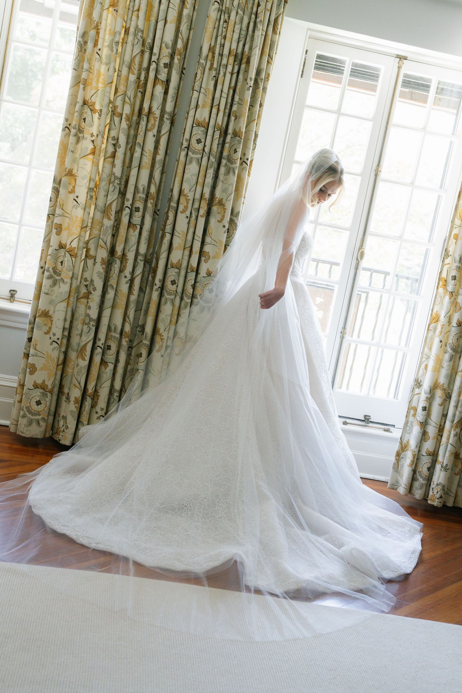Bride in a flowing white wedding gown with long veil stands by sunlit windows with floral curtains.