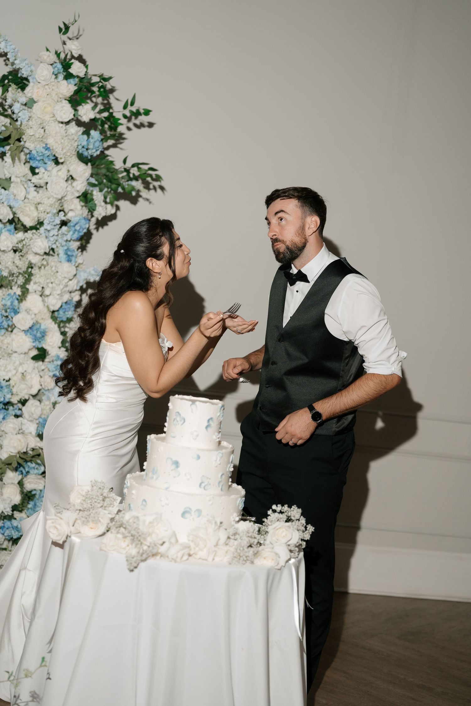 Bride and groom laugh while cutting four-tier white wedding cake adorned with blue floral decor.