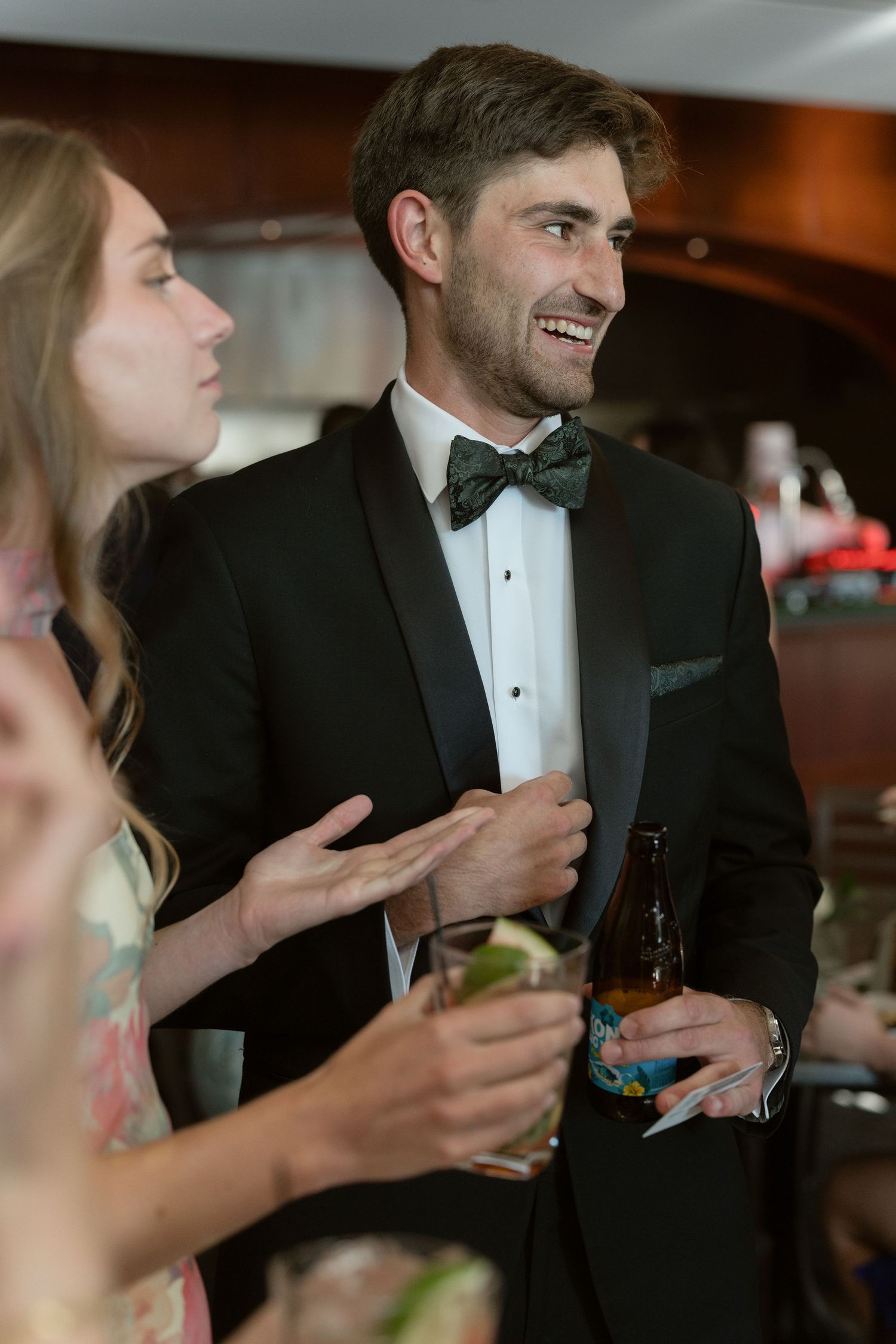 Smiling groomsman in black tuxedo and bow tie holds a beer while chatting with guests at a wedding reception.