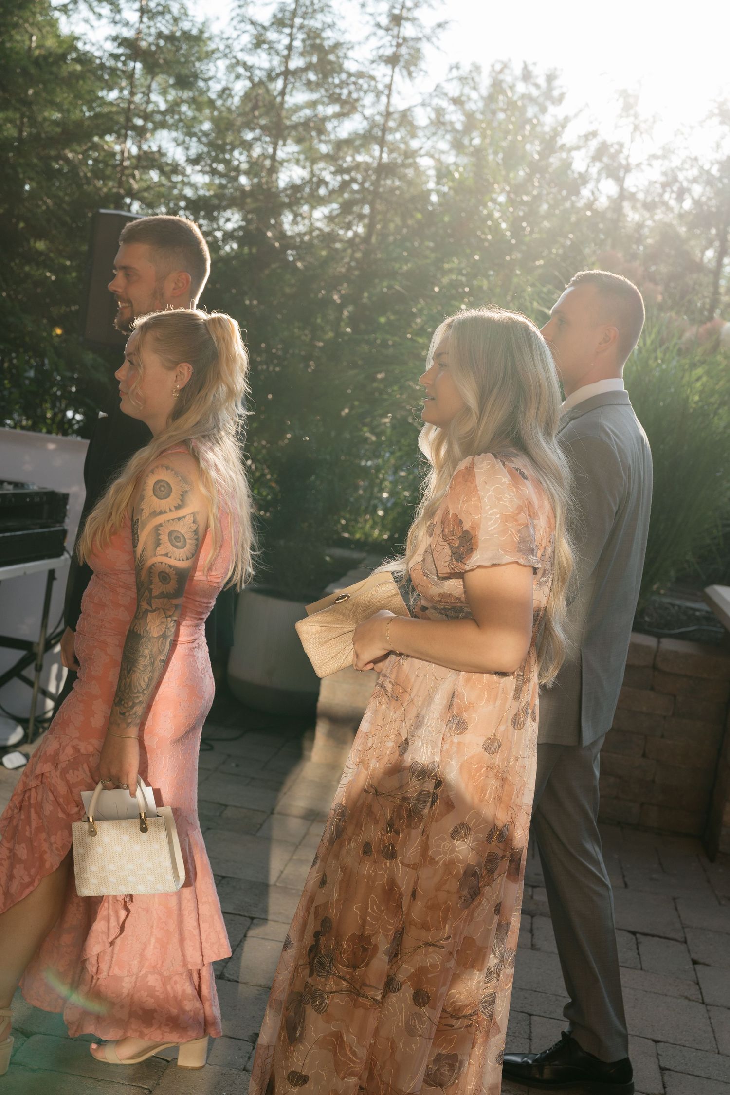 Four elegantly dressed wedding guests standing outdoors in golden sunlight, women wearing floral and pink gowns.