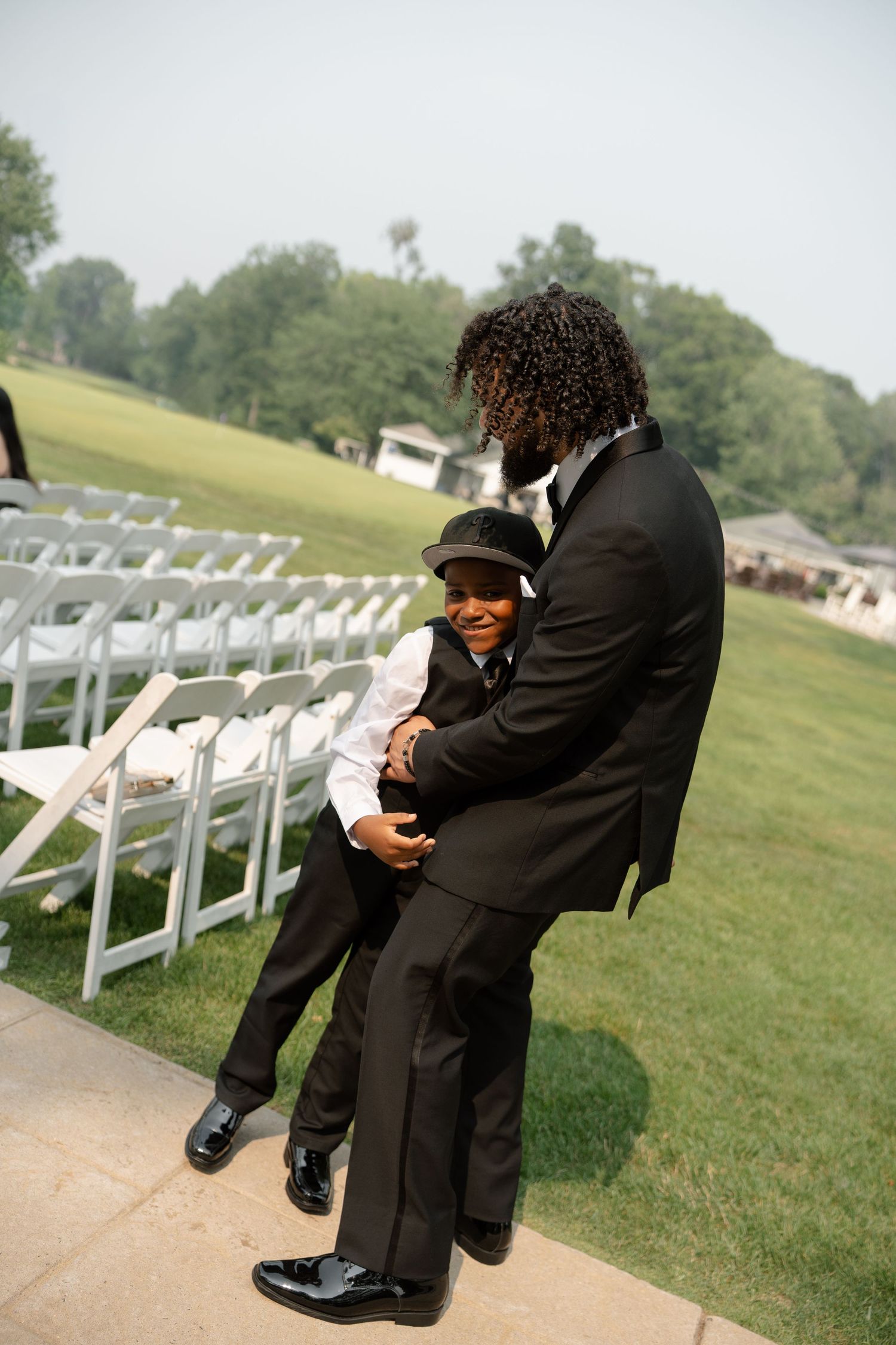 A groom in a black tuxedo shares a joyful moment with a young boy in matching attire at a wedding.