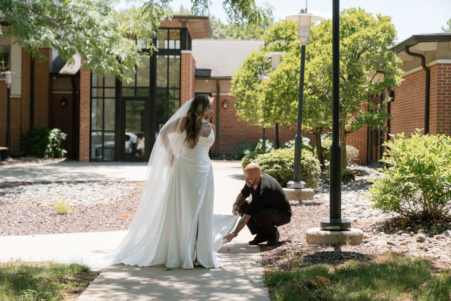 Groom kneeling to fix bride's dress on outdoor walkway near brick building on wedding day.