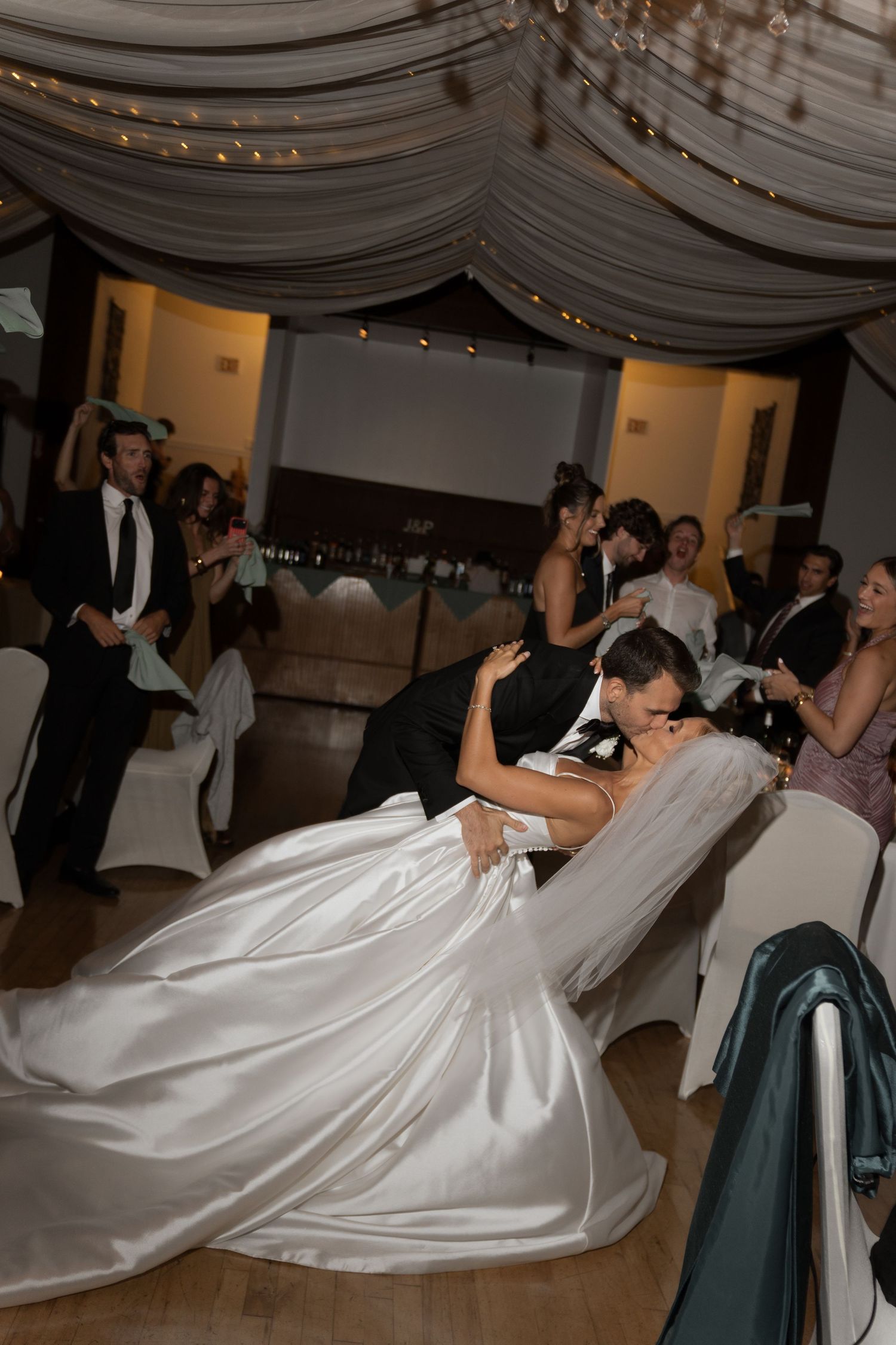 Groom dips bride during first dance at elegant wedding reception with draped ceiling and guests watching.