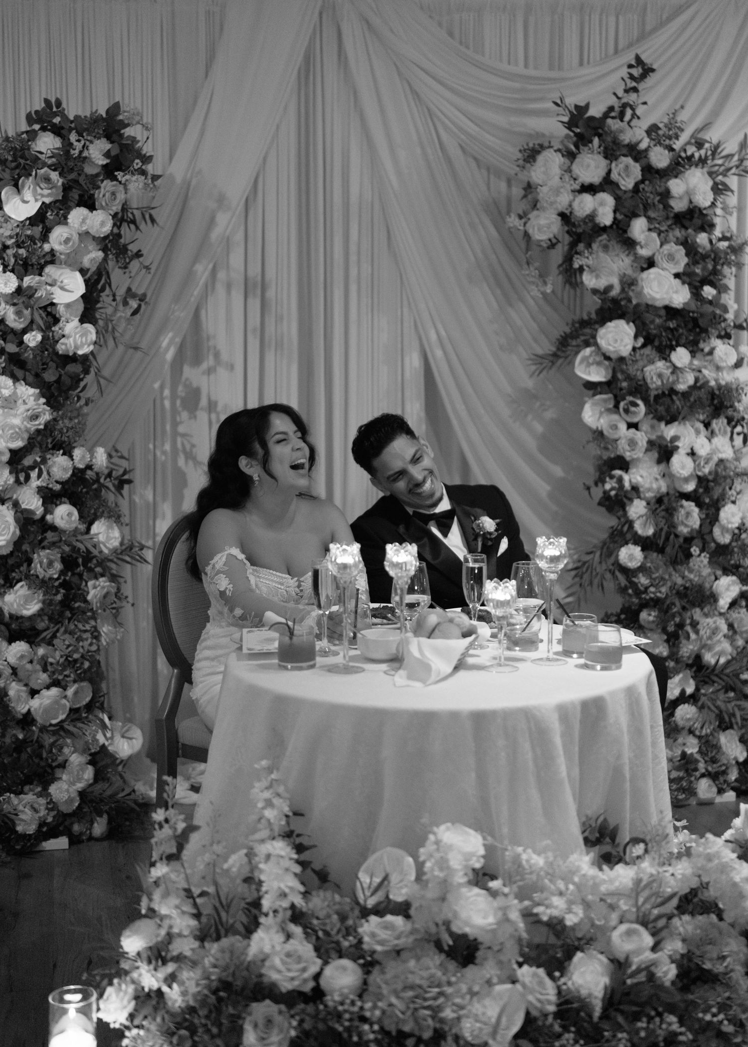 Black and white photo of newlyweds laughing at sweetheart table surrounded by lush floral arrangements.