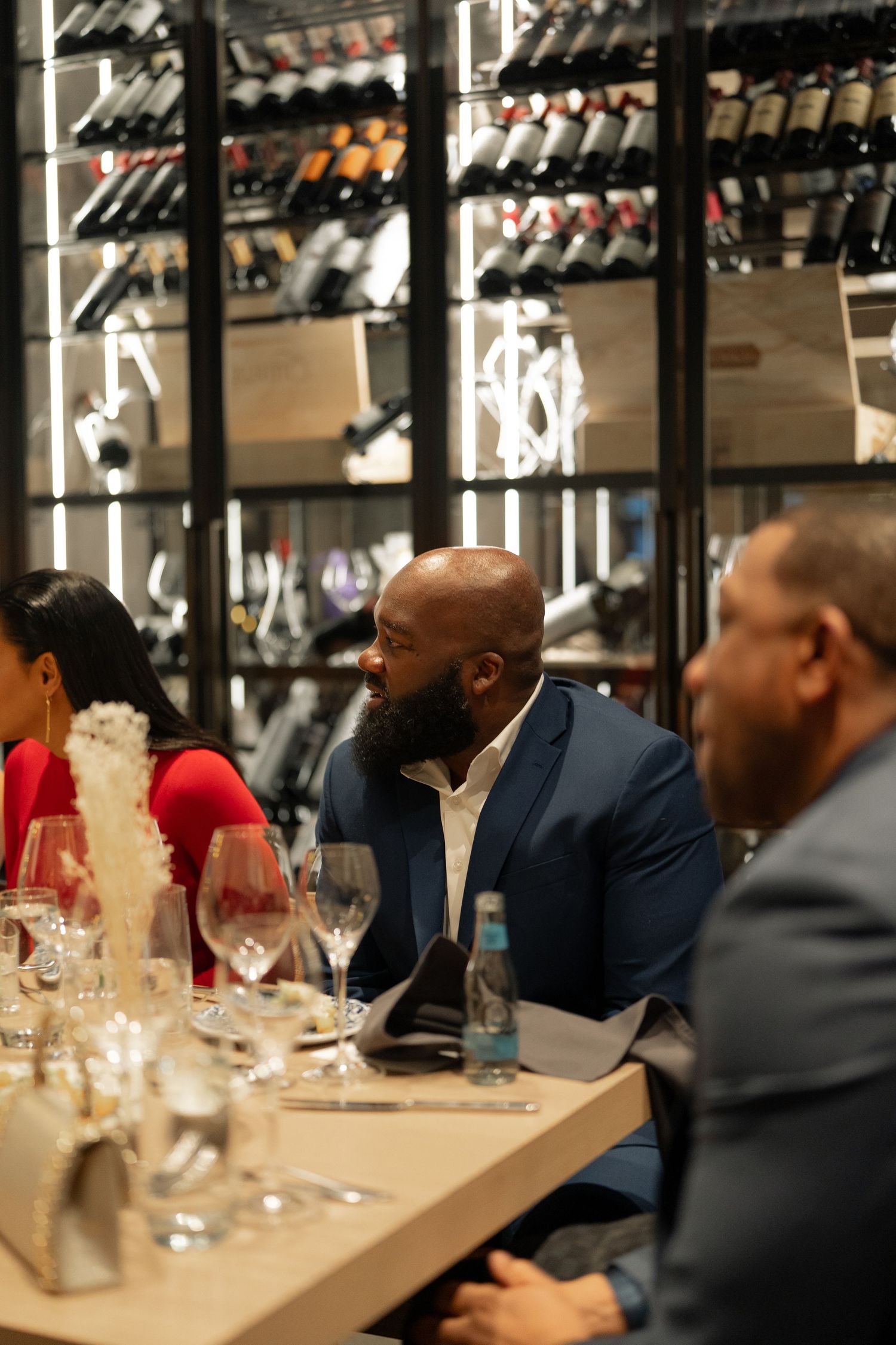 Business professionals gathered at an upscale wine bar with wine glasses and bottles displayed on shelves.
