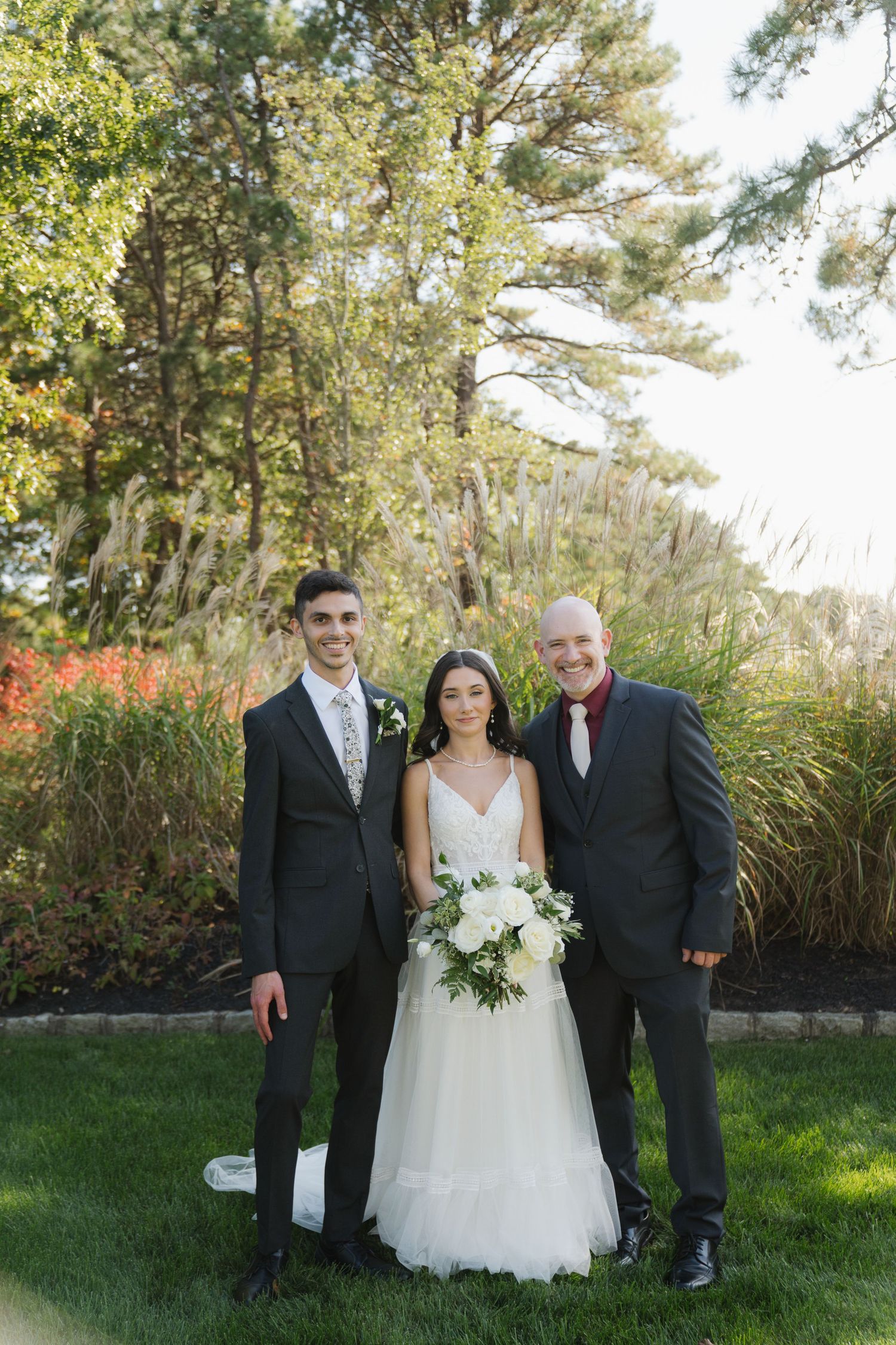 Wedding party poses together on grassy lawn with ornamental grasses and trees in the background.