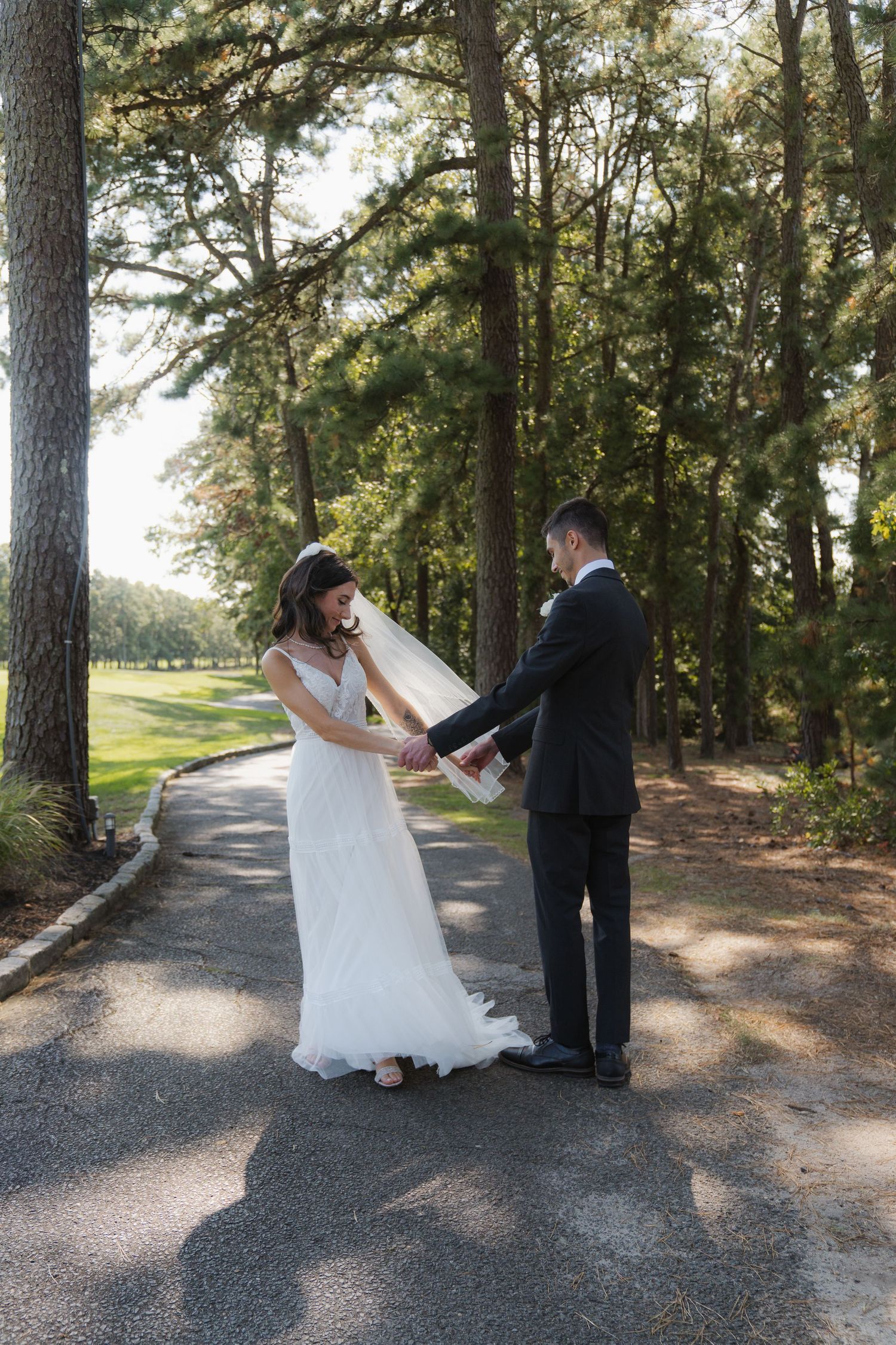 A couple shares a romantic moment on a tree-lined path during their wedding photoshoot in natural sunlight.