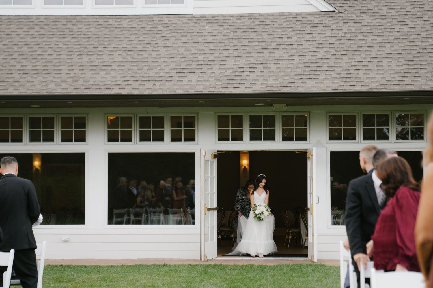 A wedding ceremony takes place on a green lawn in front of a white building with colonial-style windows.